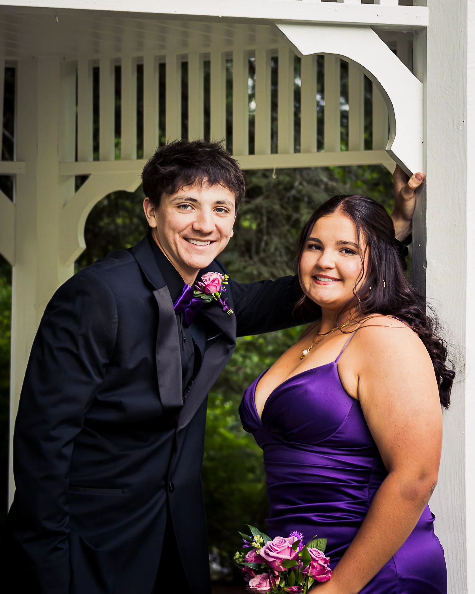 A young man and woman dressed formally, smiling outdoors at a wedding, with the man holding a bouquet of pink and purple flowers.
