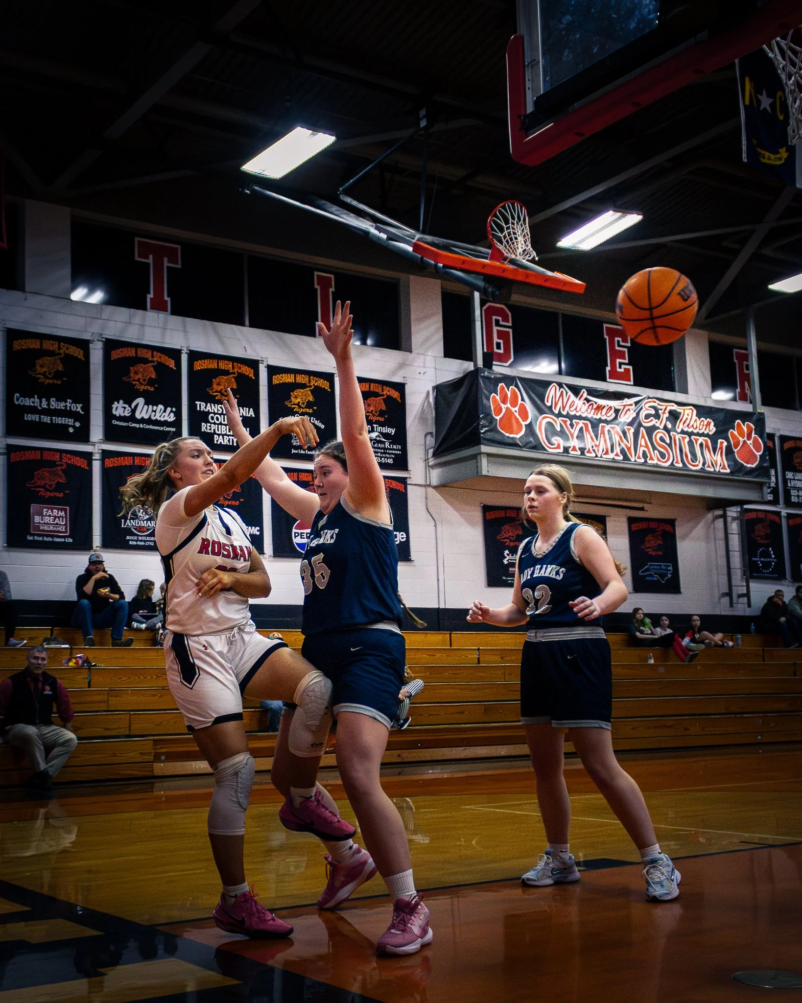 Three female basketball players on the court during a game, with one attempting a shot and two defending, inside a gymnasium with banners and a scoreboard in the background.