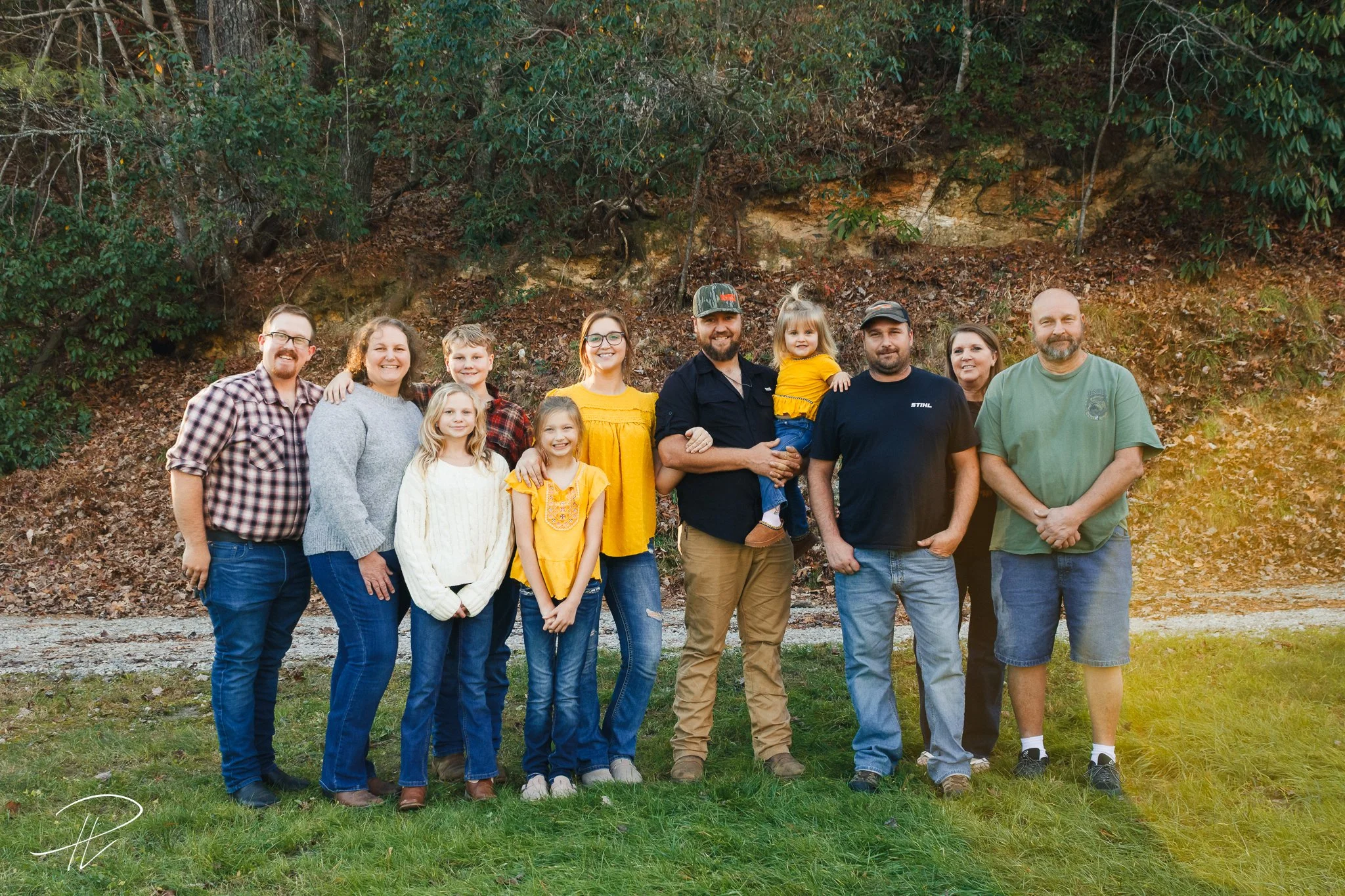 A large family poses for a photo outdoors, standing on grass with a rocky hillside and trees in the background, during late afternoon with sunlight filtering in.