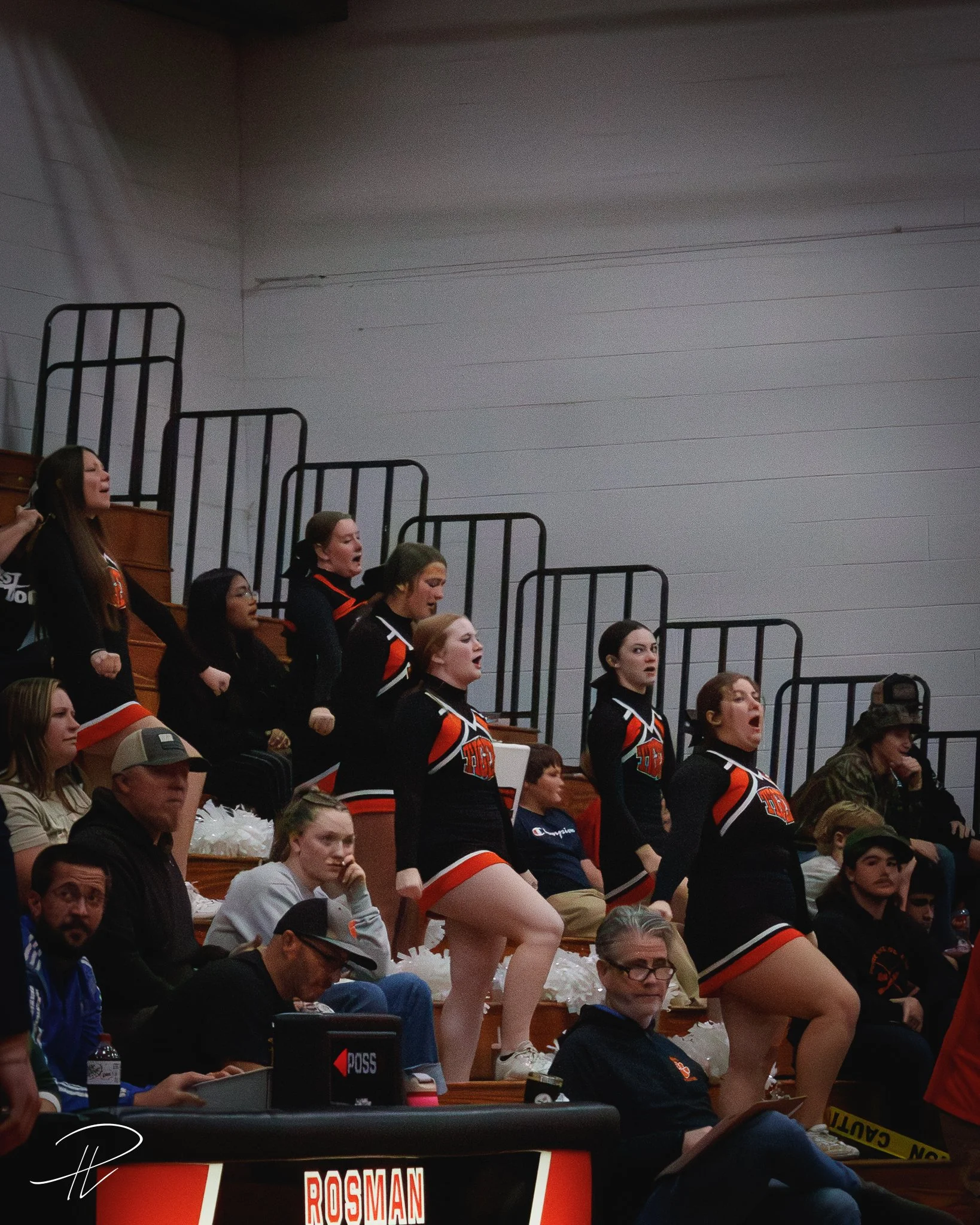 Cheerleaders performing on the bleachers at an indoor sports event, dressed in black and orange uniforms, with spectators watching.