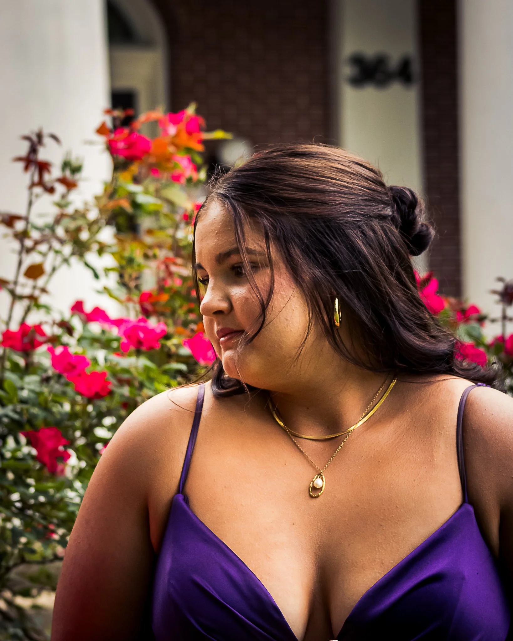 A woman with dark brown hair styled in loose waves and a braided section, wearing gold jewelry including a necklace and earrings, dressed in a purple satin dress with spaghetti straps, standing outdoors amidst pink and red flowering plants.