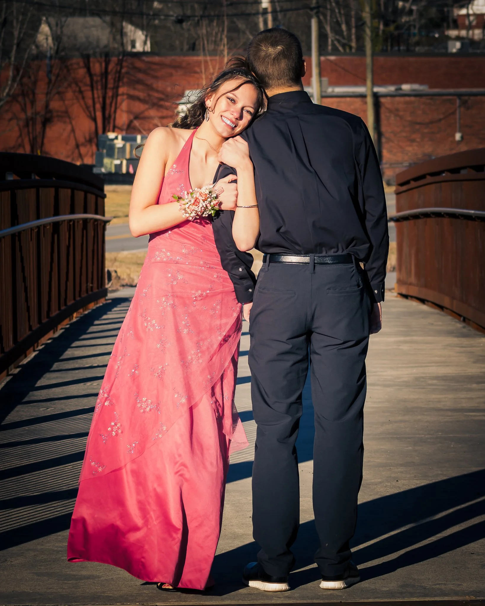 A young woman in a pink dress and a corsage smiling and leaning on a young man in a dark shirt and pants, standing on a bridge outdoors.