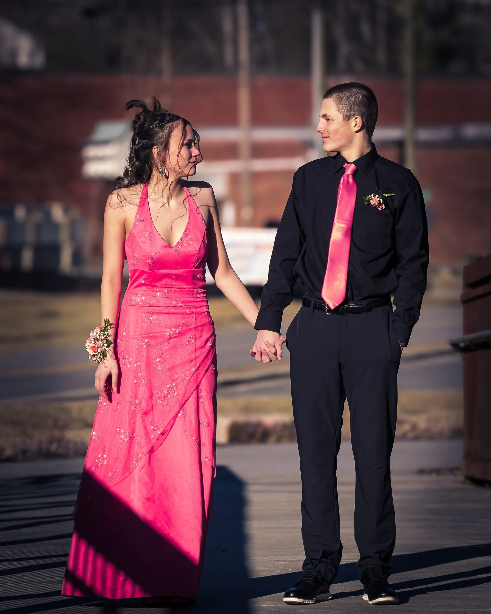 A young couple holding hands, dressed formal, walking outdoors on a sunny day. The woman is wearing a pink gown with floral details and the man is in a black shirt with a pink tie and boutonniere.