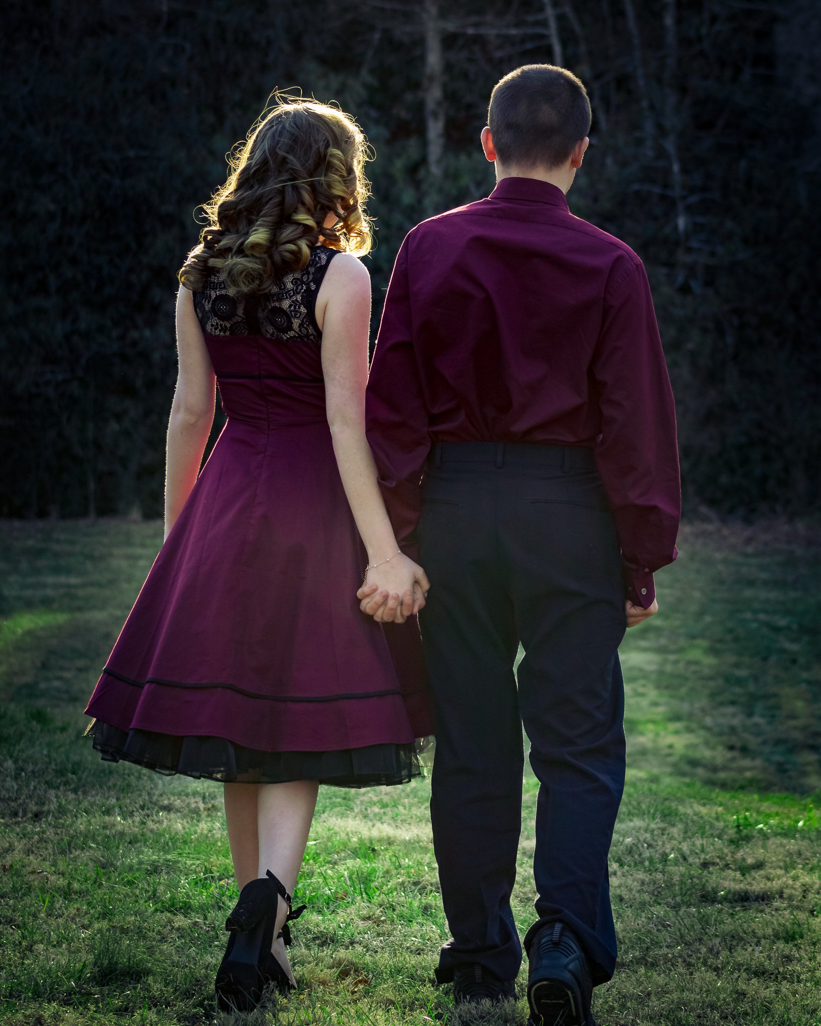 A couple holding hands and walking on grass outdoors during sunset, seen from behind.