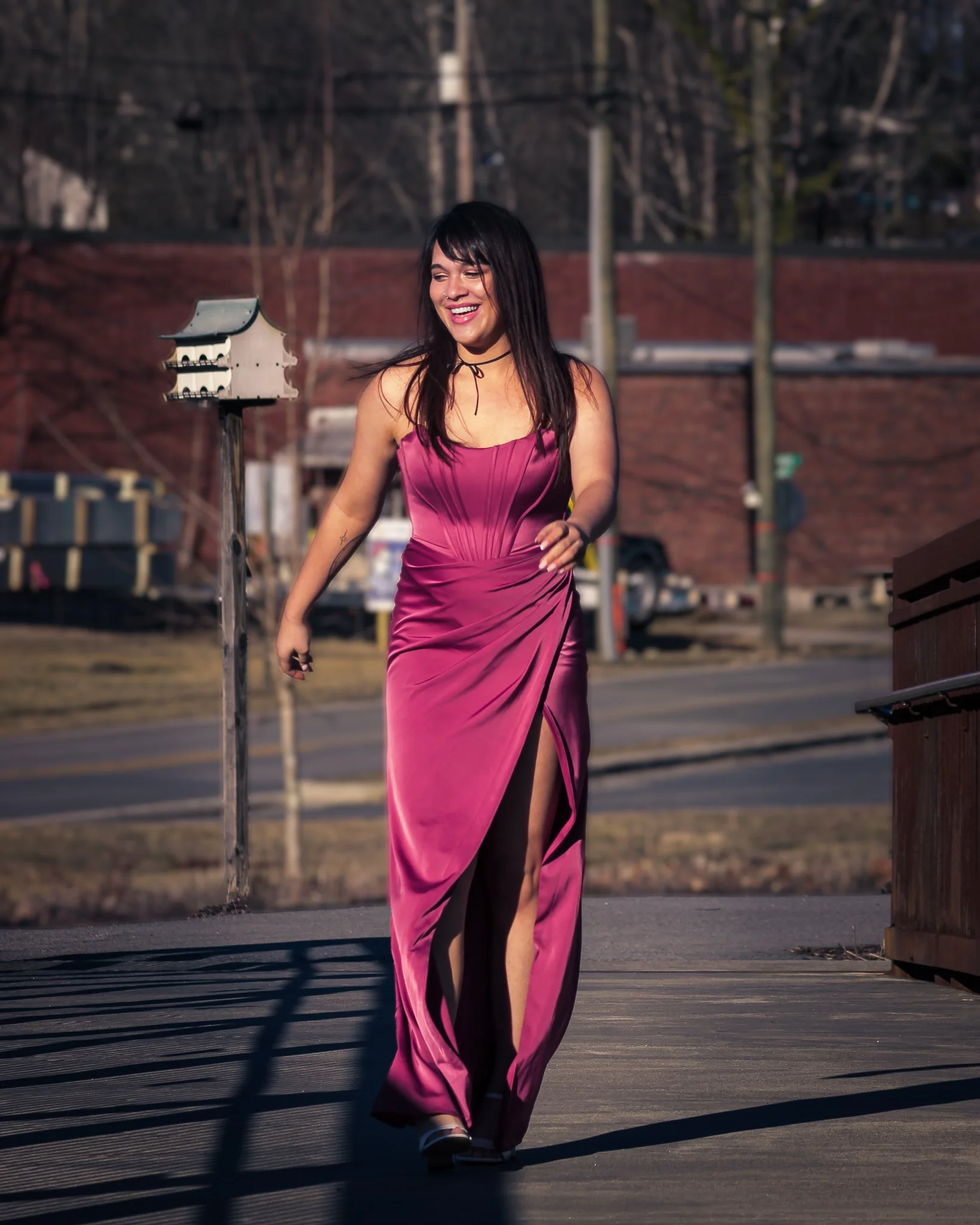 A woman in a long, pink satin dress with a front slit, walking outdoors on a sidewalk, smiling.