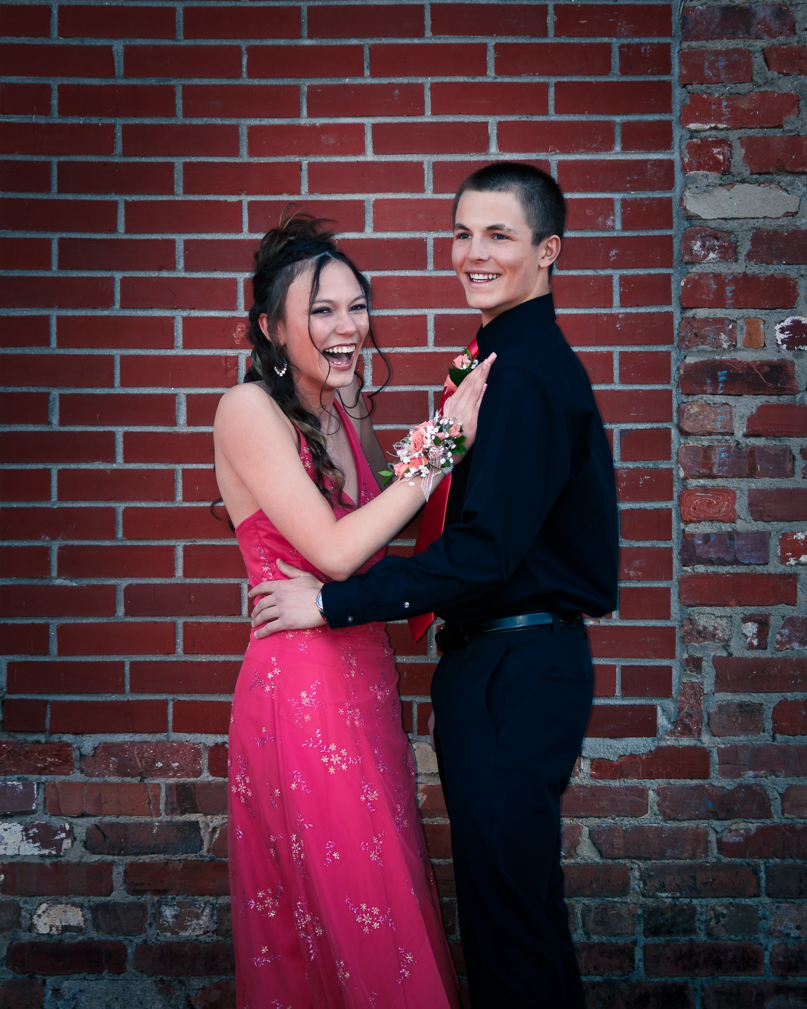 A young couple standing in front of a red brick wall, dressed formally, with the girl in a pink dress and the boy in a black shirt and pants. They are smiling and holding each other, with the girl holding a corsage.