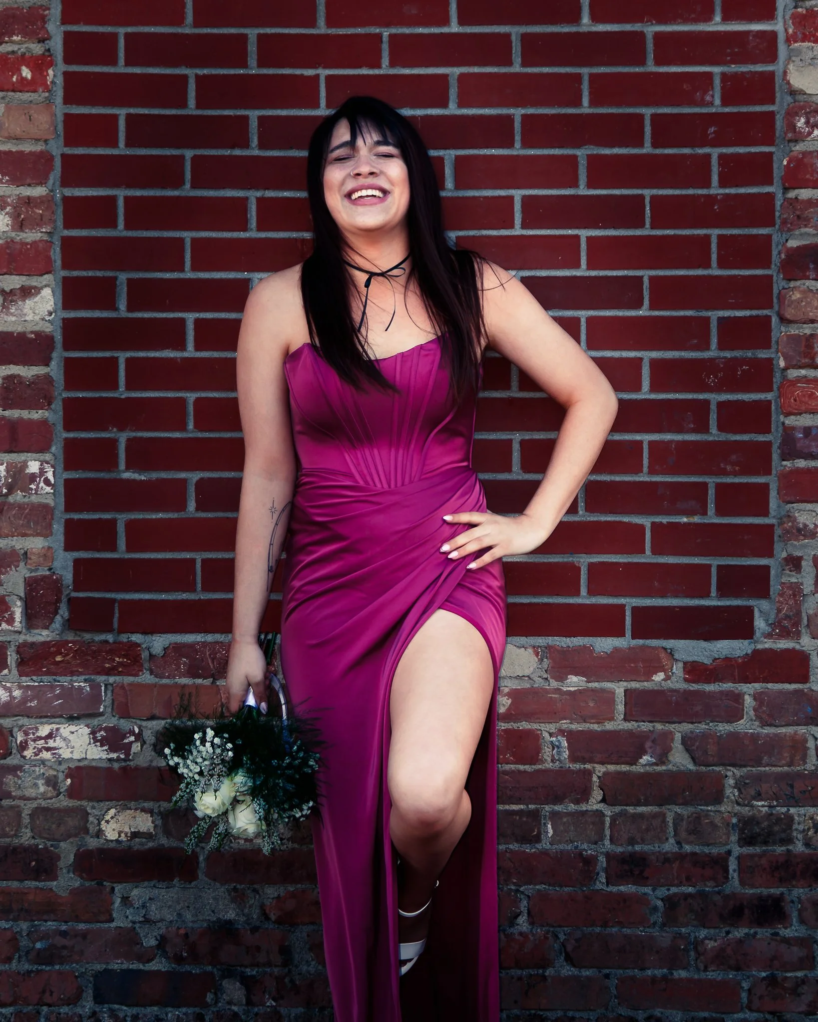 A young woman in a magenta dress with a high slit, holding a bouquet of white roses and greenery, standing against a brick wall, smiling with one hand on her hip.