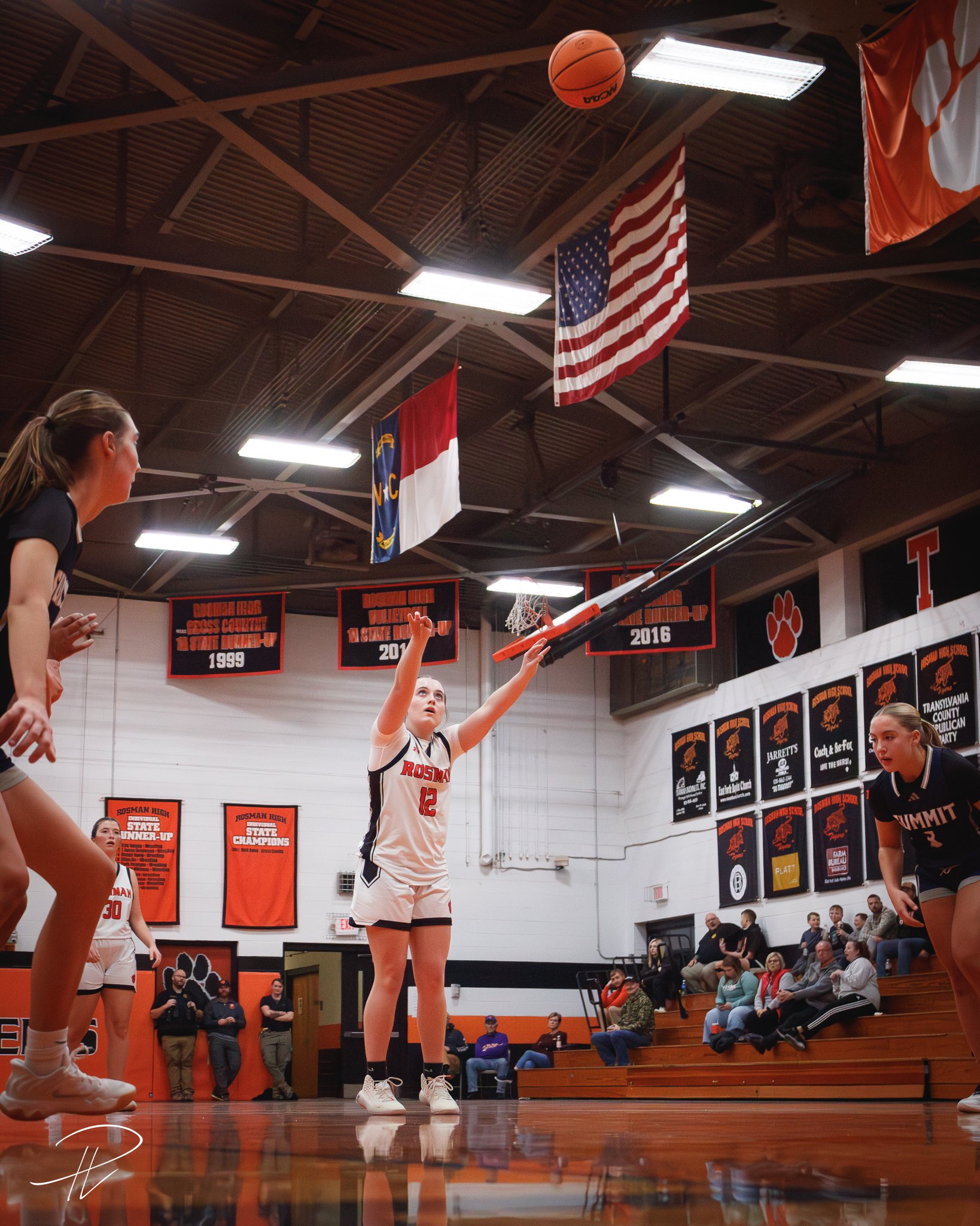 A girls' basketball game in progress inside a gymnasium, with a player shooting a basketball towards the hoop, while other players and spectators watch.