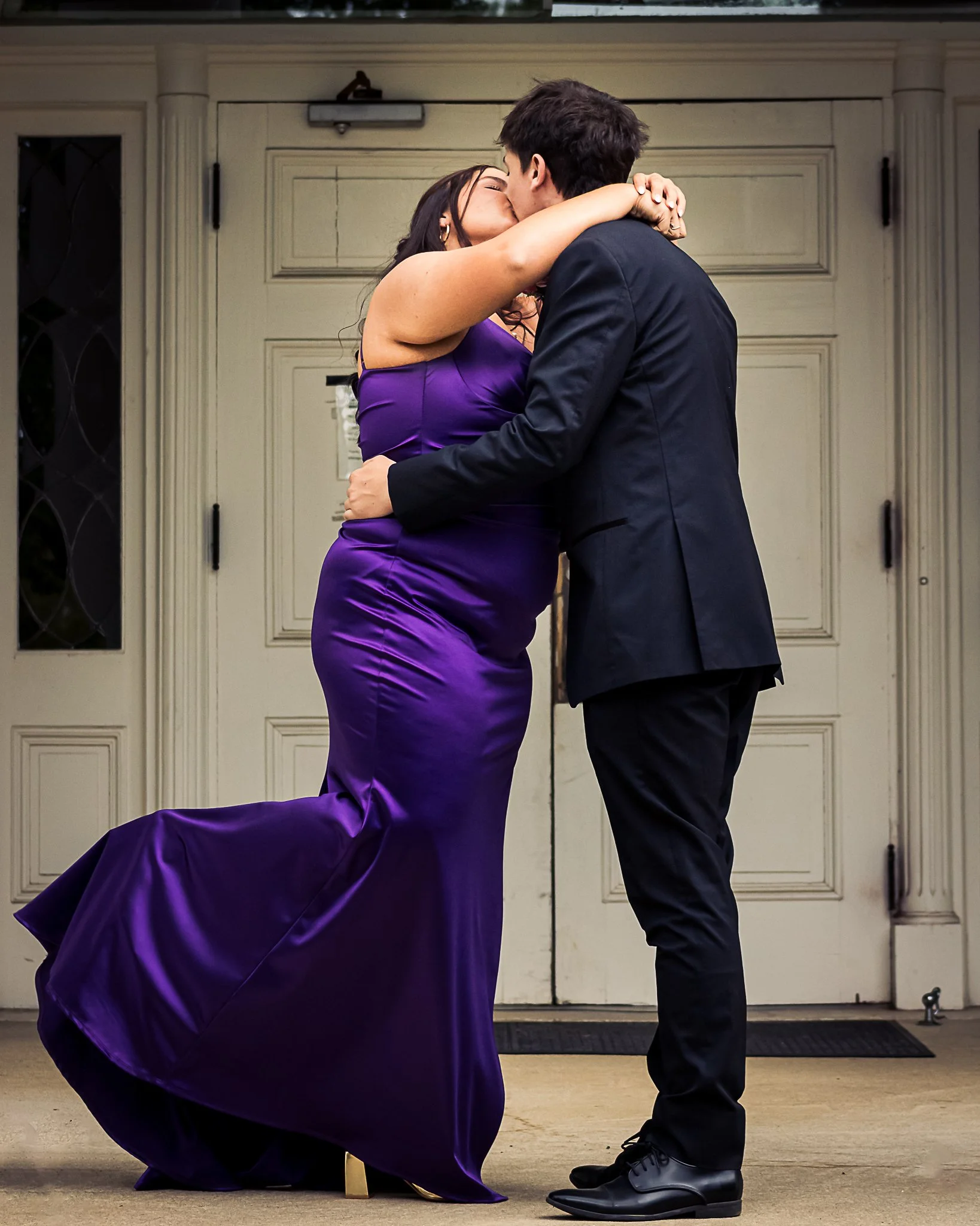 A woman in a purple dress and a man in a black suit are kissing in front of a white garage door. The woman is kneeling, and the man is standing. They are embracing each other.