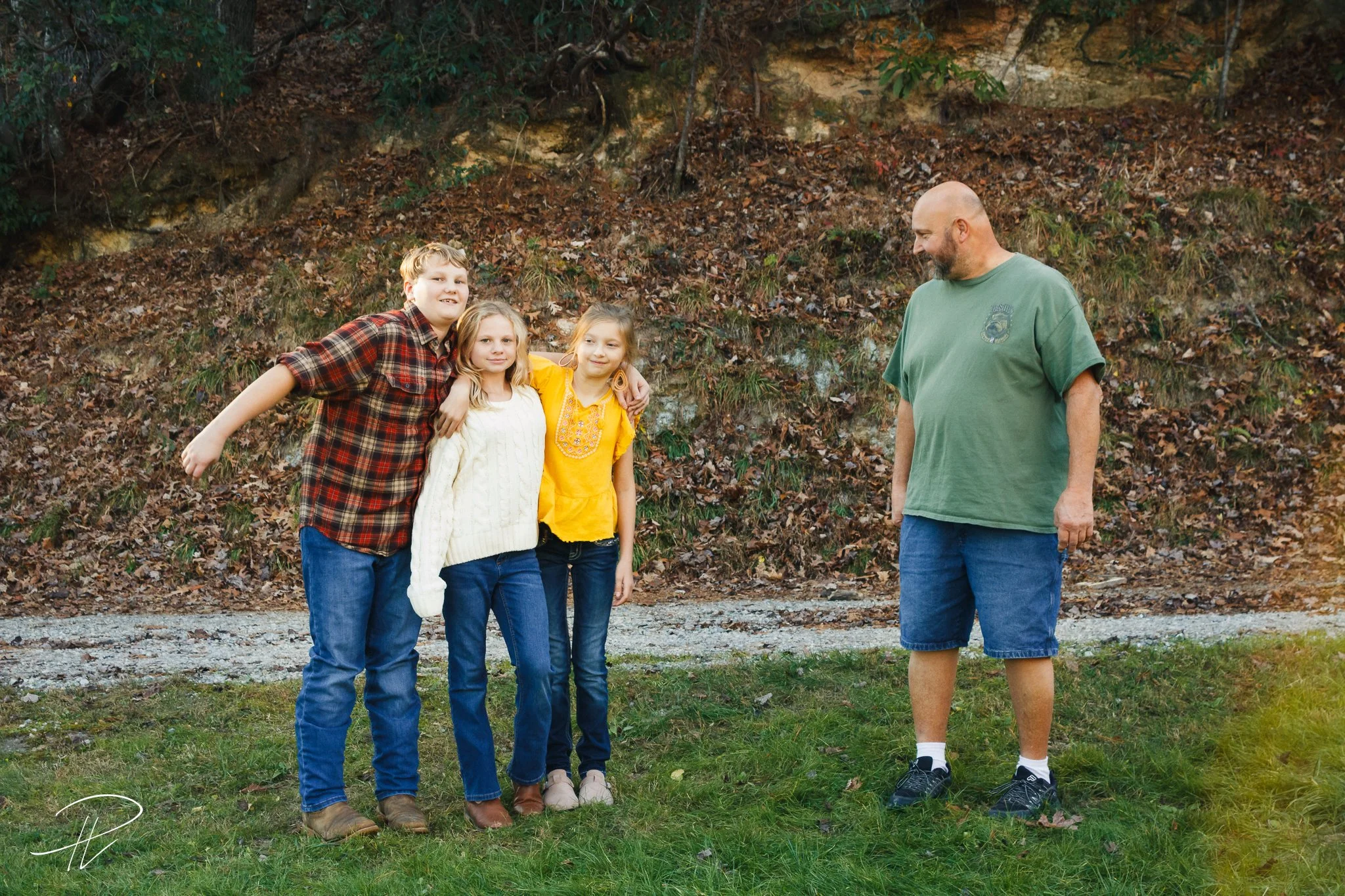 A man and three children standing outdoors on grass, with a rocky hillside in the background. The man is looking at the children, who are standing close together, smiling.