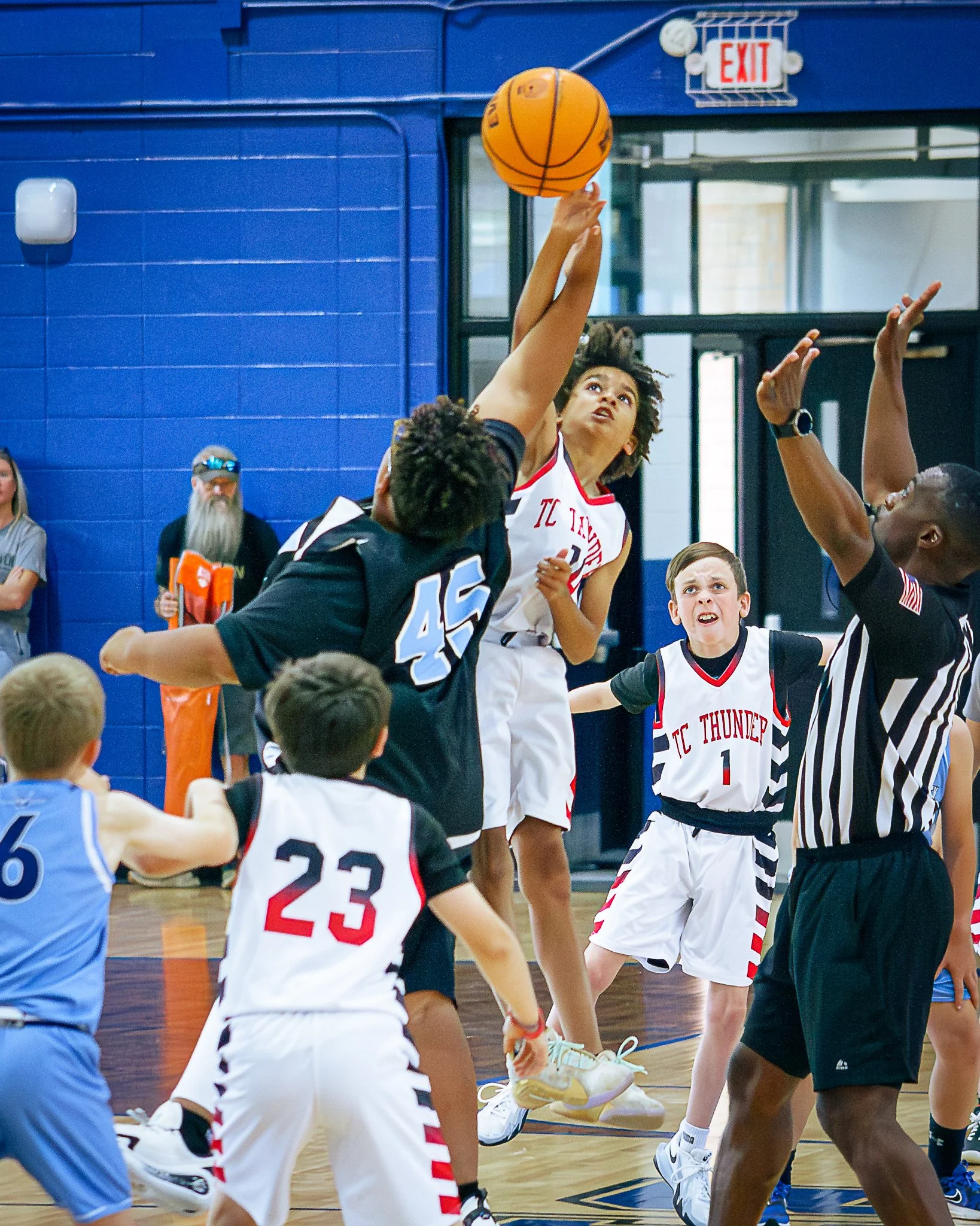 Children playing basketball in an indoor gym, reaching for the ball, with a woman referee overseeing the game. Spectators watch from the sidelines.