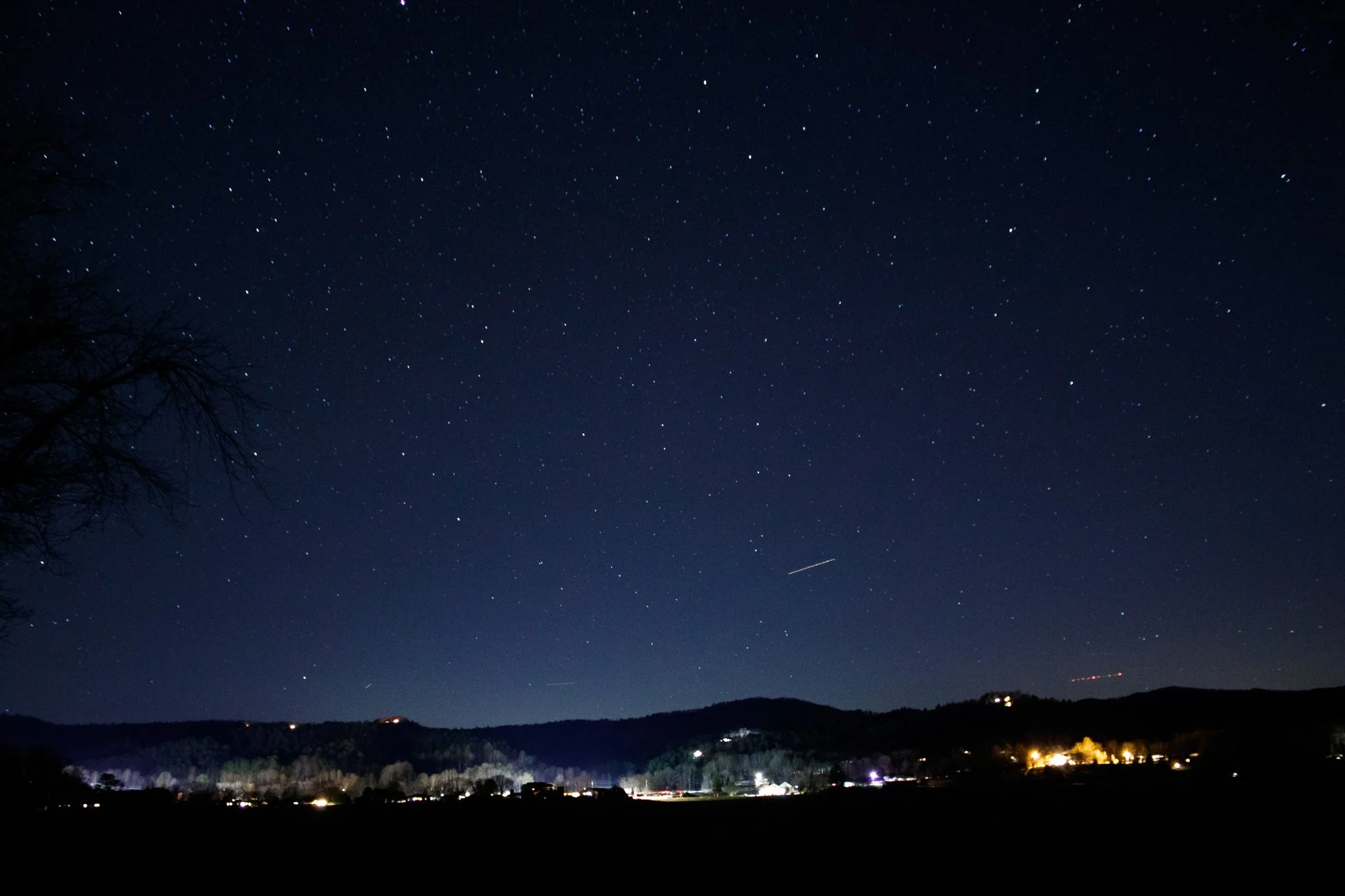 Night sky filled with stars over a landscape with hills and scattered lights from houses or buildings.