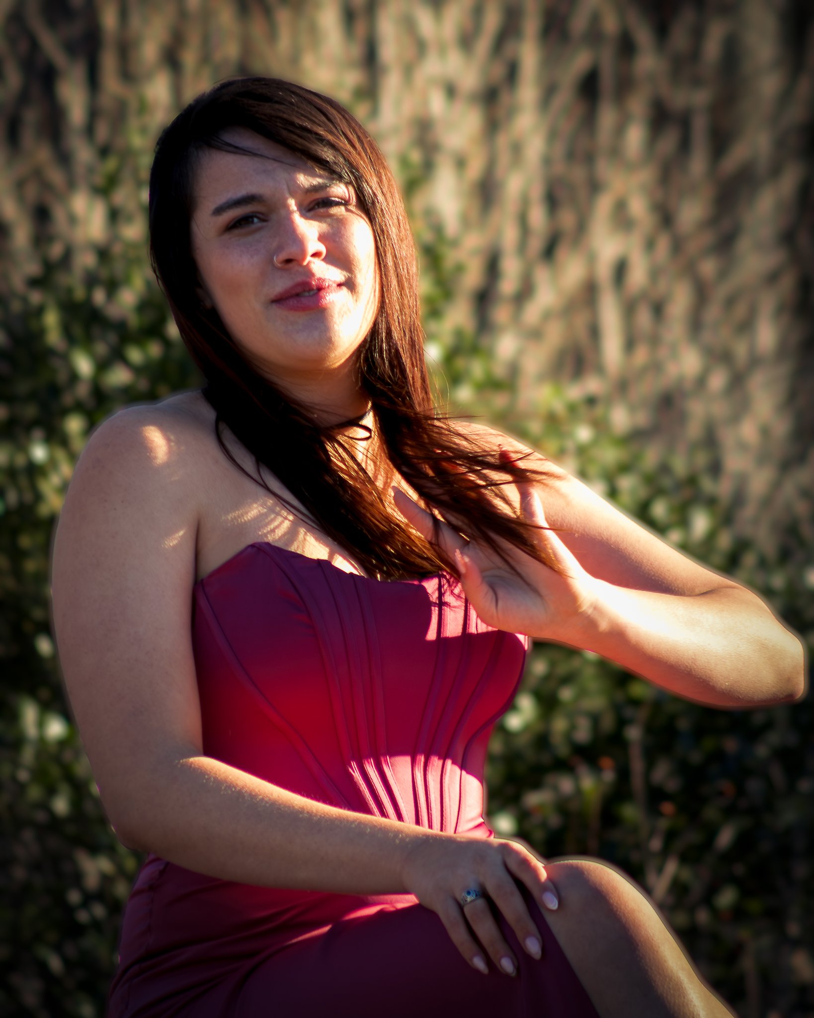 A woman in a strapless burgundy dress sitting outdoors with her hand on her chest, detailed by sunlight and a blurred background of trees.