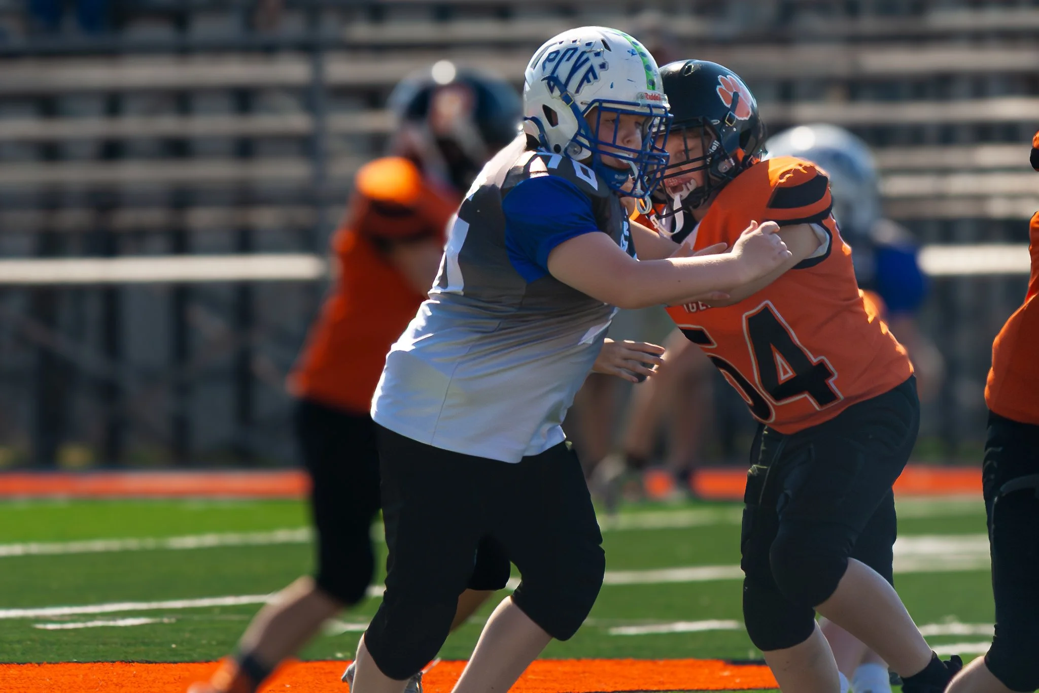 Two young football players wearing helmets and uniforms are engaged in a tackle during a game on a football field. One player is in a blue and white uniform, while the other is in an orange and black uniform. The players are in close contact, with th