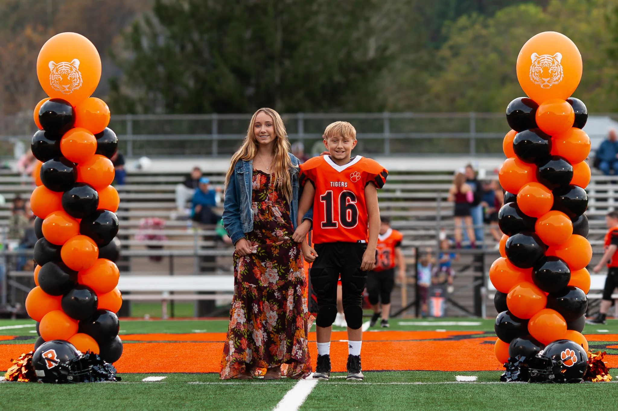 A boy in a football uniform and a girl in a dress stand under decorated balloon columns with a tiger logo during a football game.