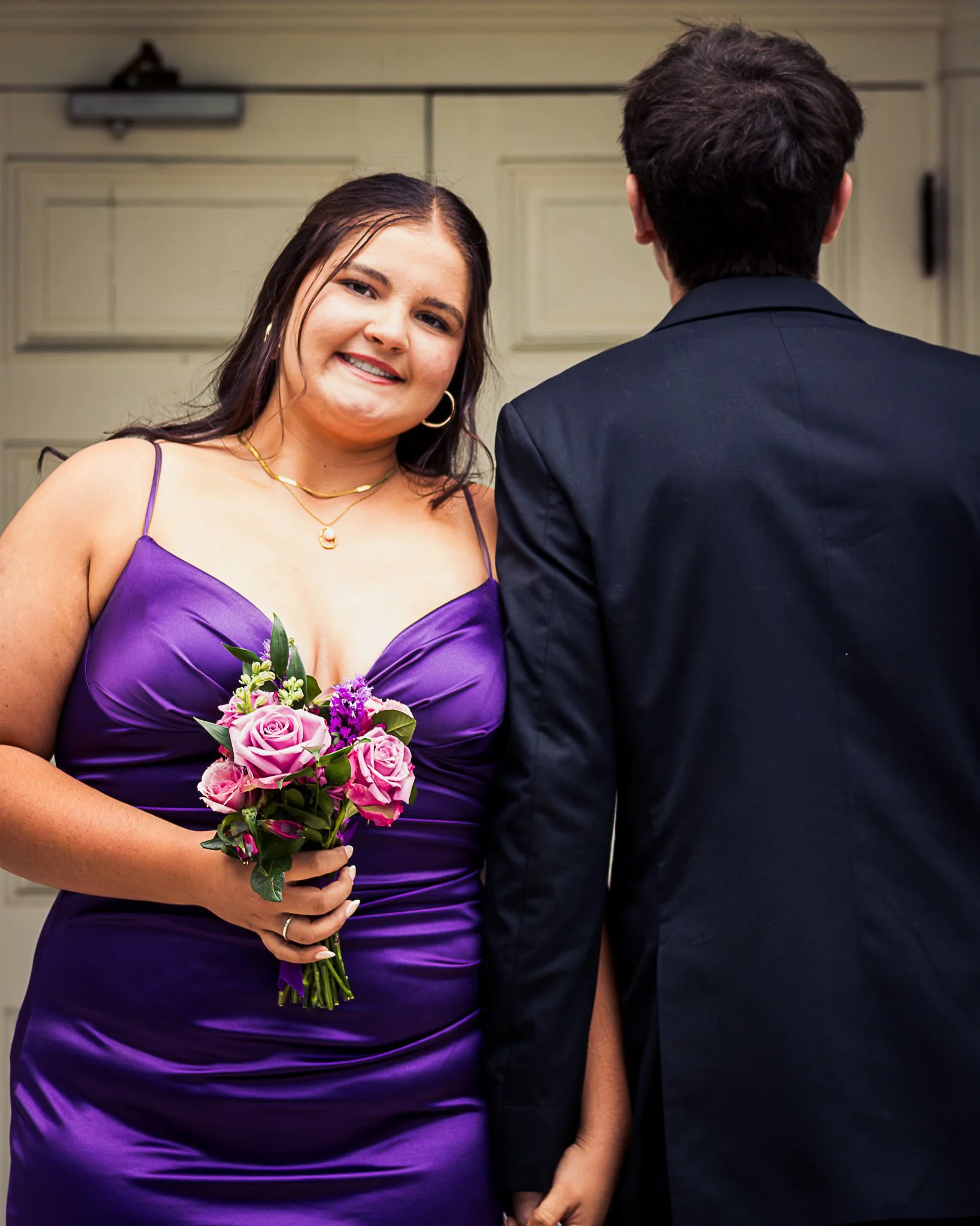 A woman in a purple dress holding a bouquet of pink and purple flowers, standing next to a man in a dark suit. She is smiling and looking at the camera.