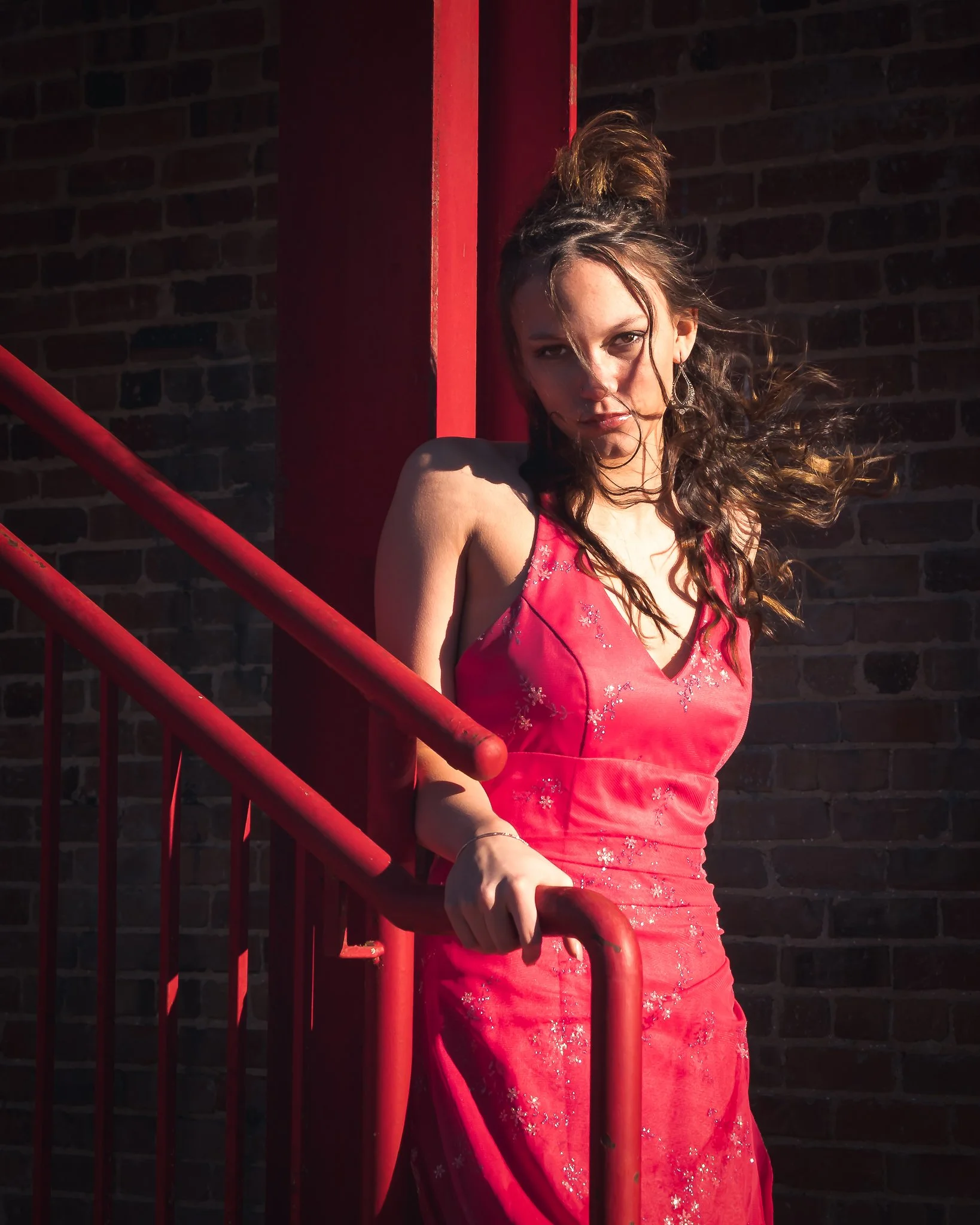 A woman in a pink dress standing next to red metal railing outside against a brick wall, with wind blowing her hair.