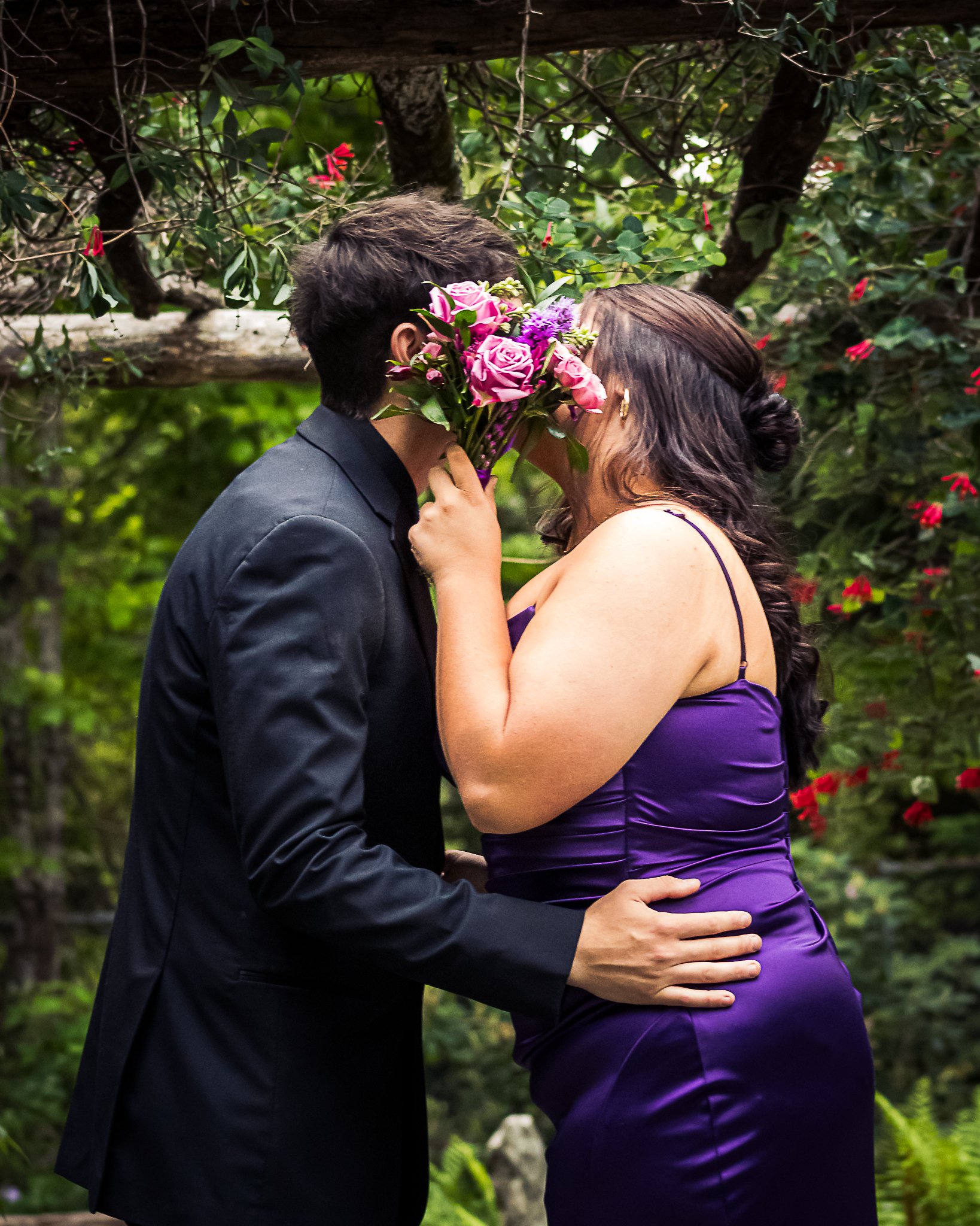 A couple is romantically kissing outdoors, the woman is holding a pink bouquet in front of her face, and they are surrounded by green foliage and pink flowers.