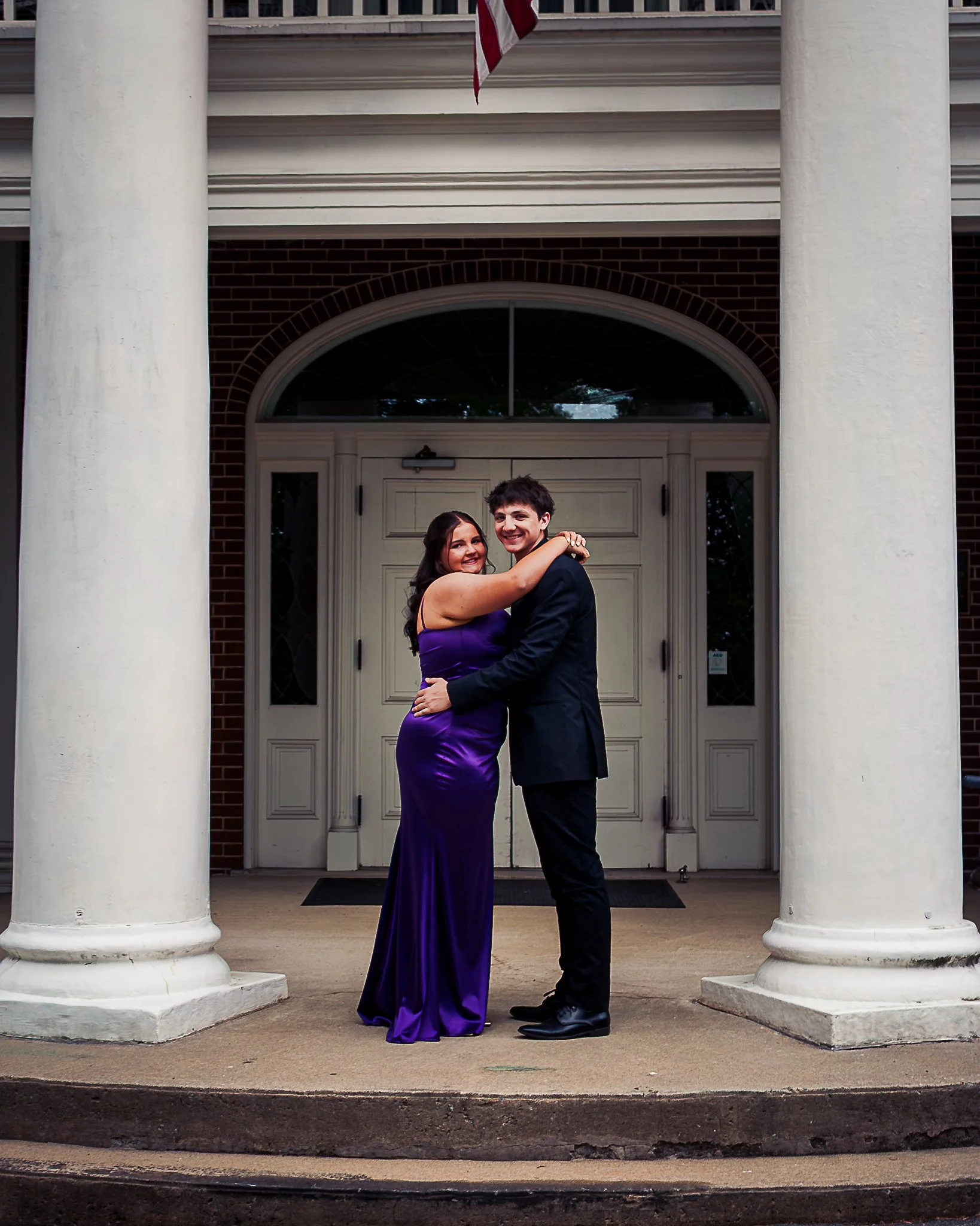 A young couple, a woman in a purple dress and a man in a suit, standing on the front steps of a building with large white columns, embracing each other and smiling.