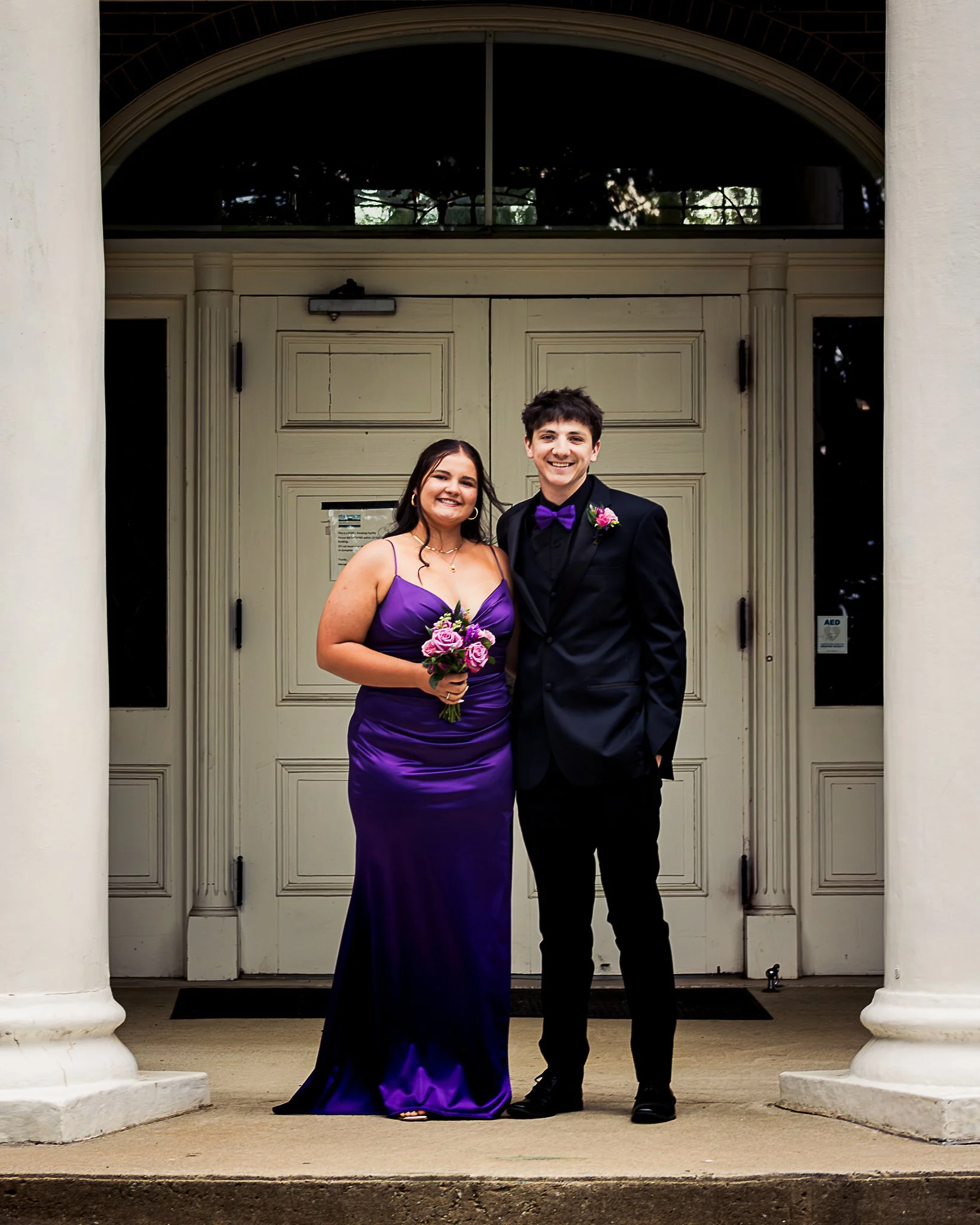 A couple standing in front of a large white door with columns on each side. The woman is wearing a purple gown and holding a bouquet of purple and pink flowers. The man is in a black tuxedo with a purple bow tie and a pink flower boutonniere. They ar