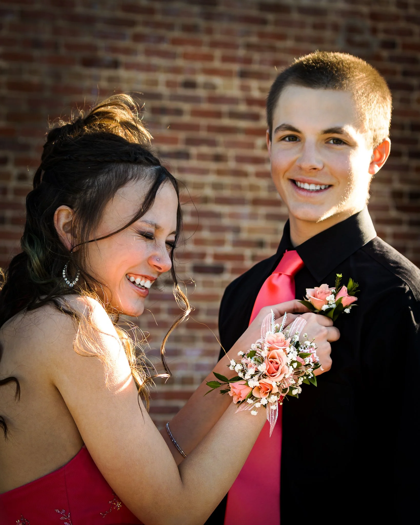A young woman and a young man dressed in formal attire, with the woman wearing a corsage and the man wearing a boutonniere, standing against a brick wall. They are smiling and looking happy.