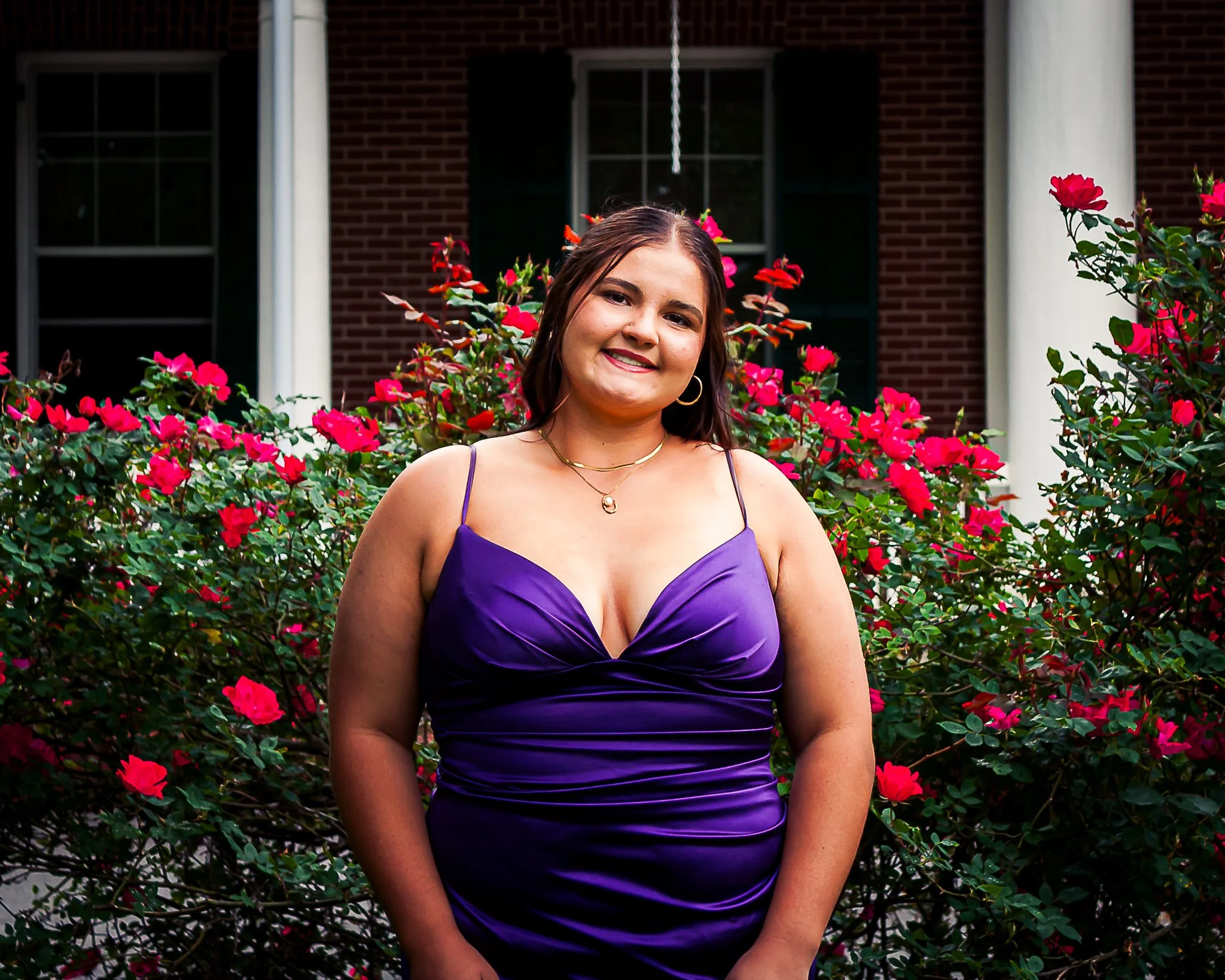 A young woman with dark hair in a purple dress standing in front of pink and red flowers outside a brick house.