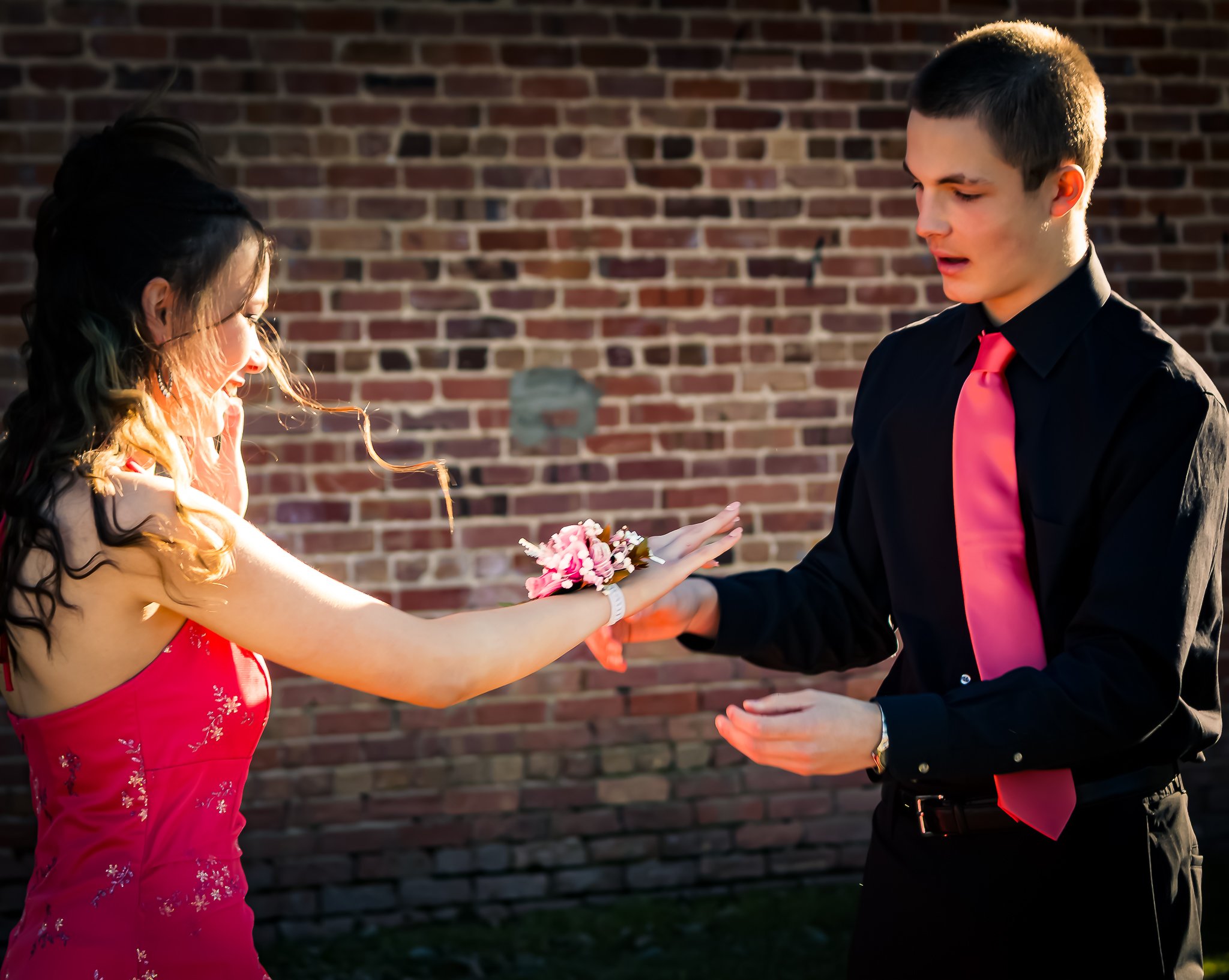 A young woman in a red dress receives a corsage from a man in a black shirt with a pink tie, standing in front of a brick wall.