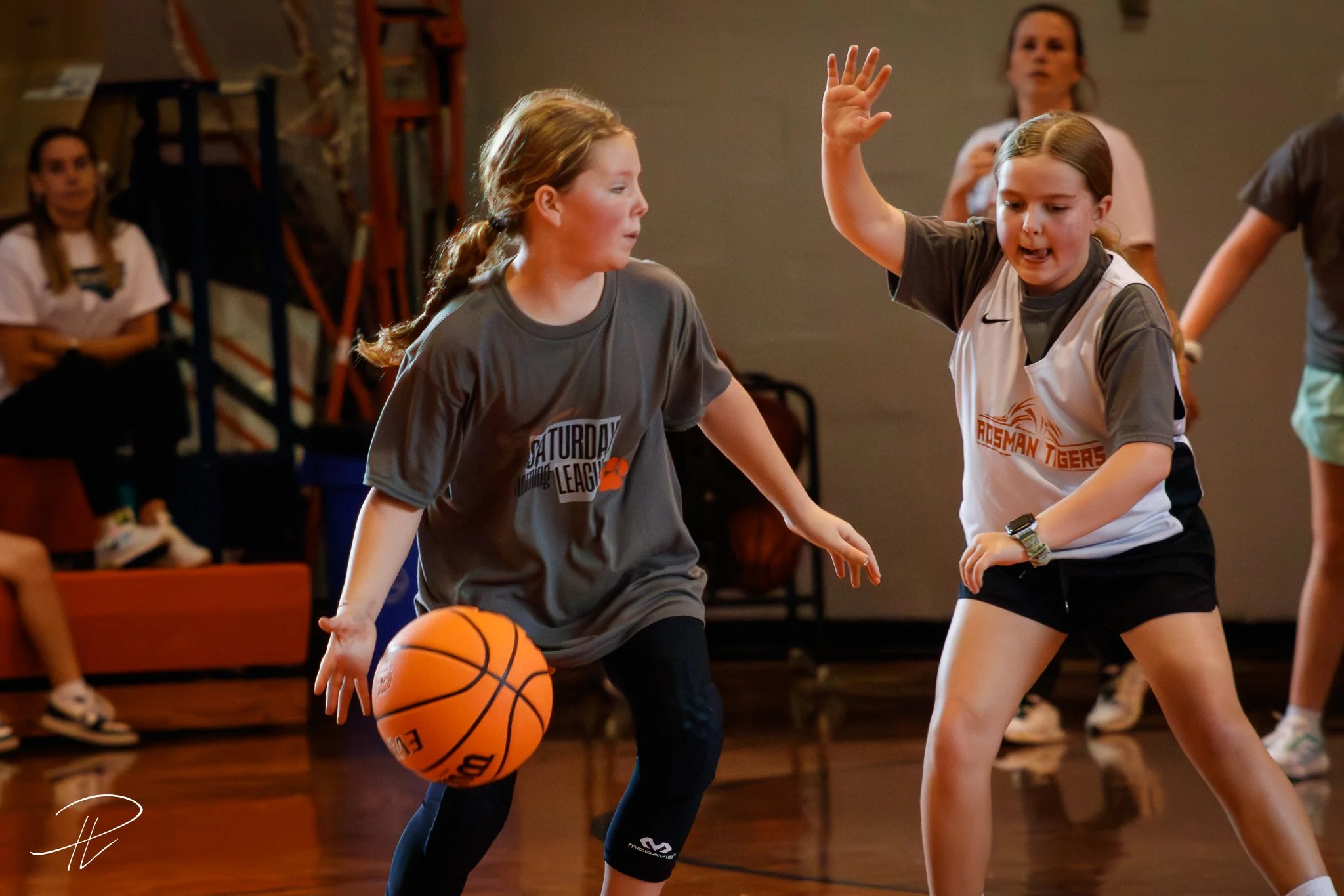Two young girls playing basketball indoors, one dribbling the ball while the other defends with her hand raised.
