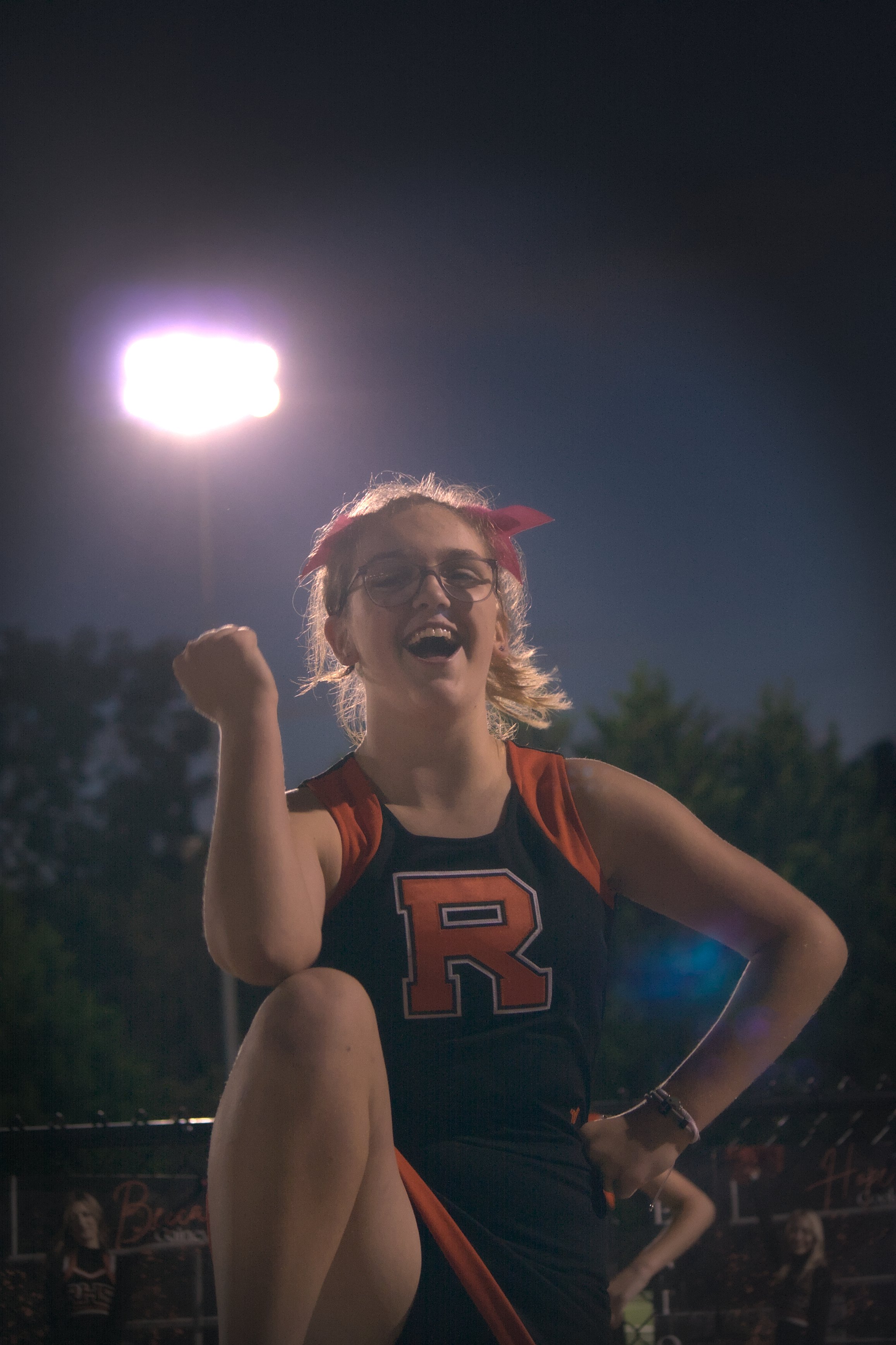 A cheerleader with glasses, blonde hair with a red bow, and a red and black uniform with an orange 'R' on the front, is smiling and raising her arm in celebration under bright stadium lights at night.