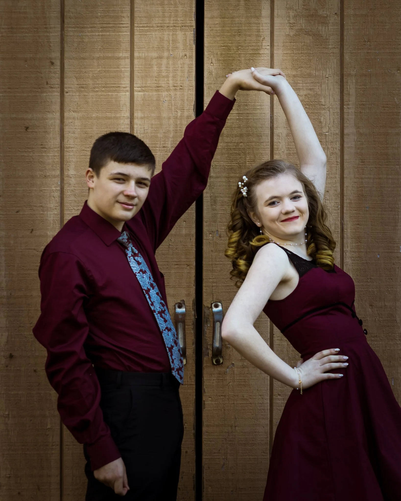 A young man and woman dance together, holding hands overhead, in front of a wooden door. They both wear matching maroon outfits, with the woman in a dress and the man in a shirt and tie.