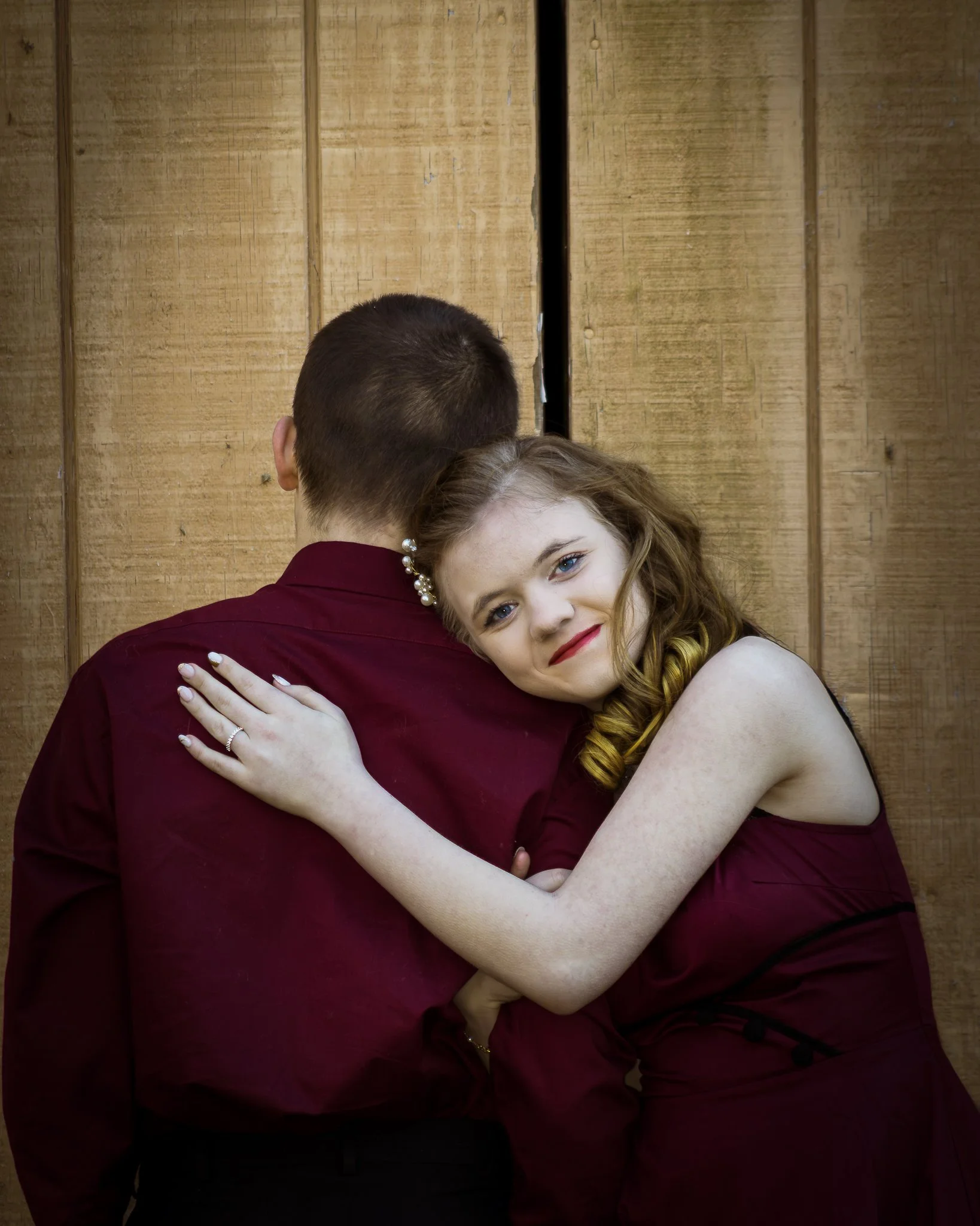 Young woman with red hair embraces a man with short hair, standing against a wooden background.