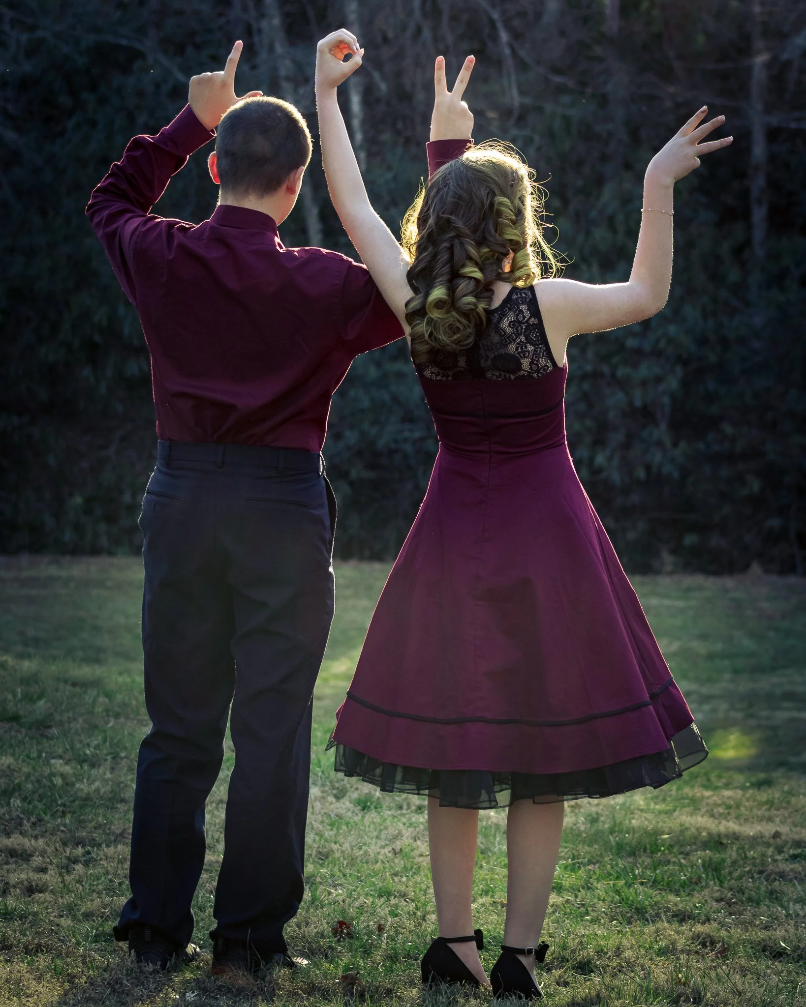A man and woman dancing together outdoors during sunset, with their backs to the camera. The woman is wearing a burgundy dress with black lace and the man is dressed in a matching burgundy shirt and black pants.