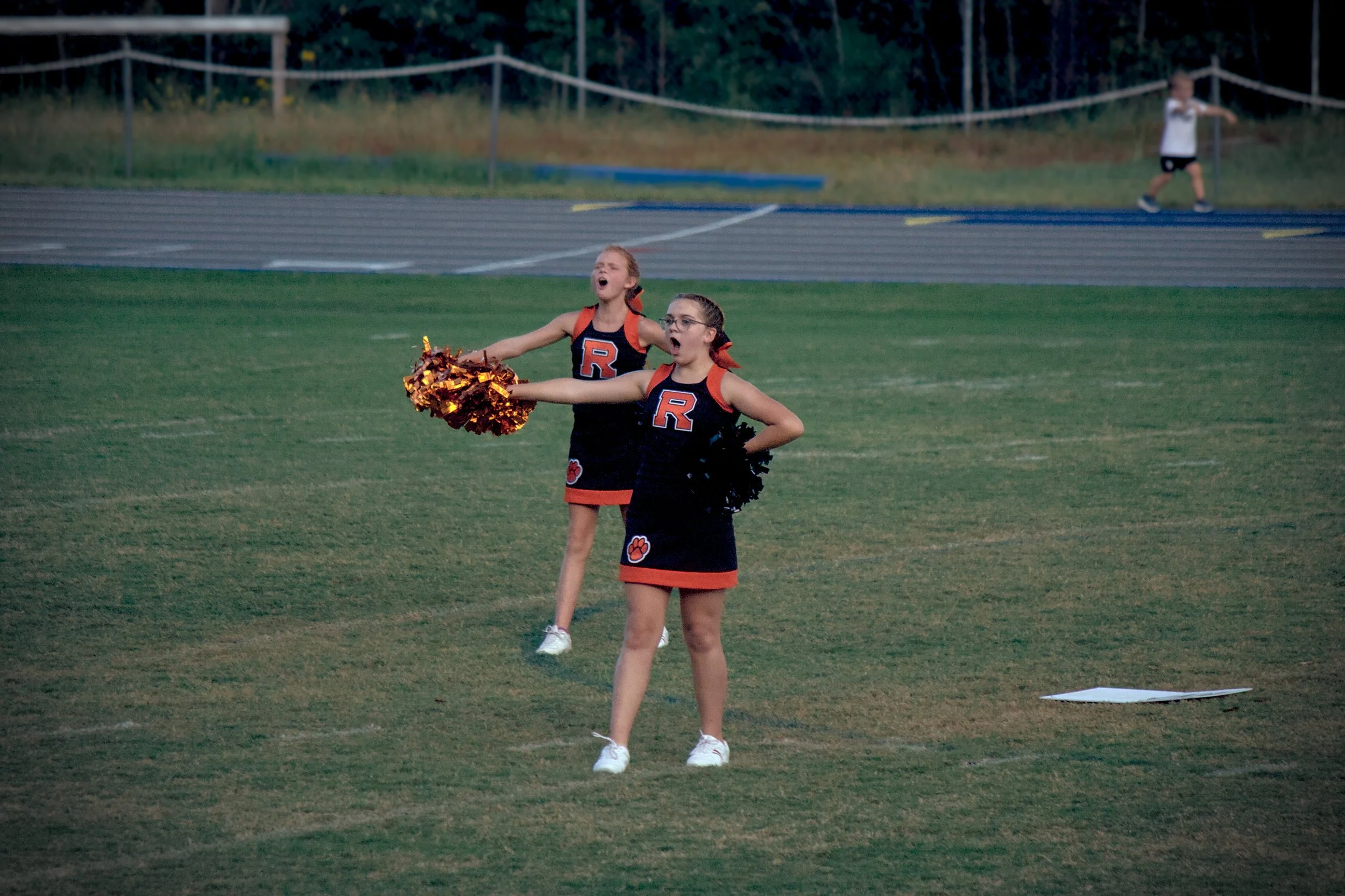 Two cheerleaders in black and orange uniforms with a large 'R' on the front perform on a sports field, holding pom-poms, with a girl in a background walking along an athletic track.