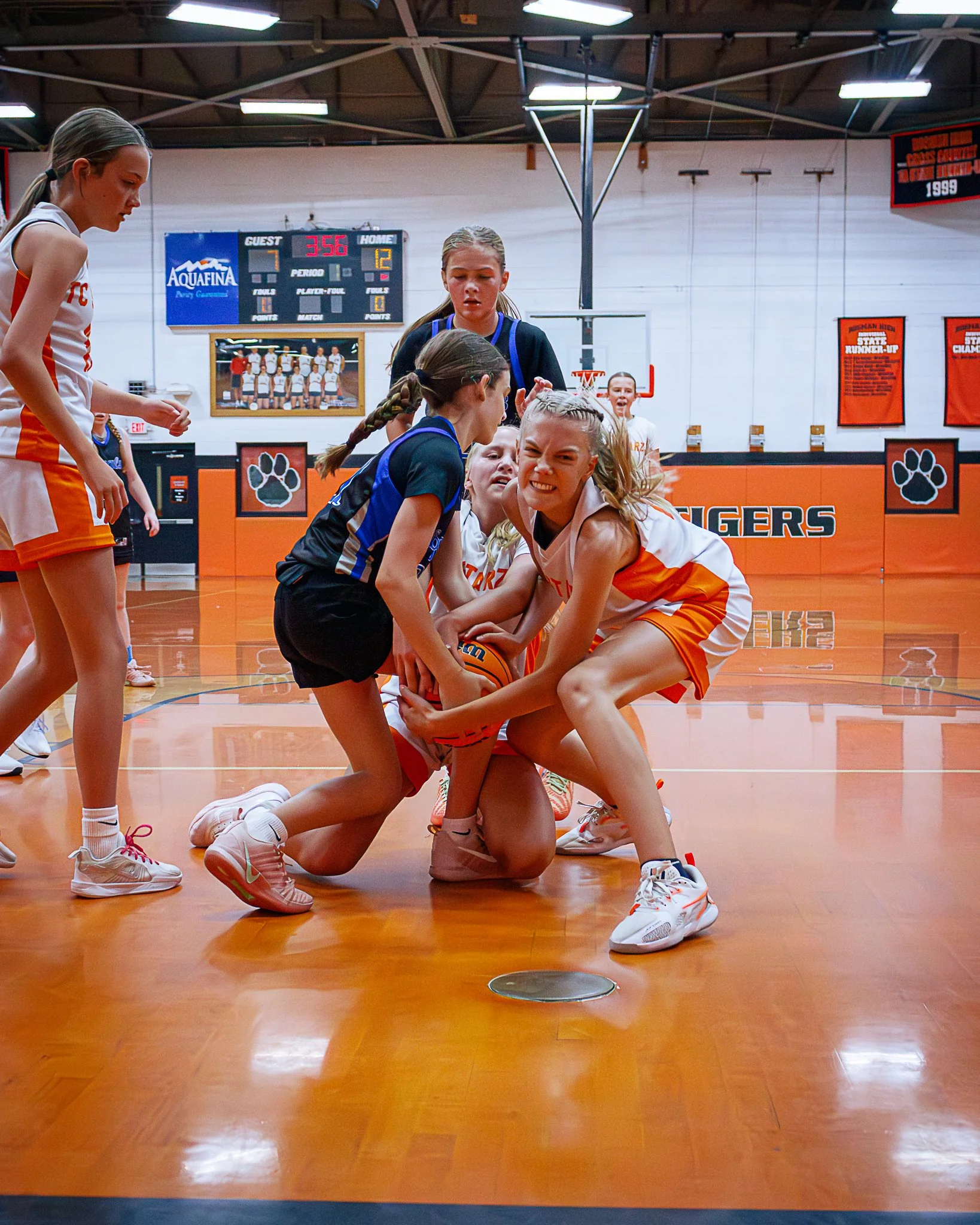 Girls' volleyball team practicing on an indoor court, some kneeling and others standing, with a scoreboard in the background showing 3:56 remaining in the game, score 12-12, and banners on the wall.