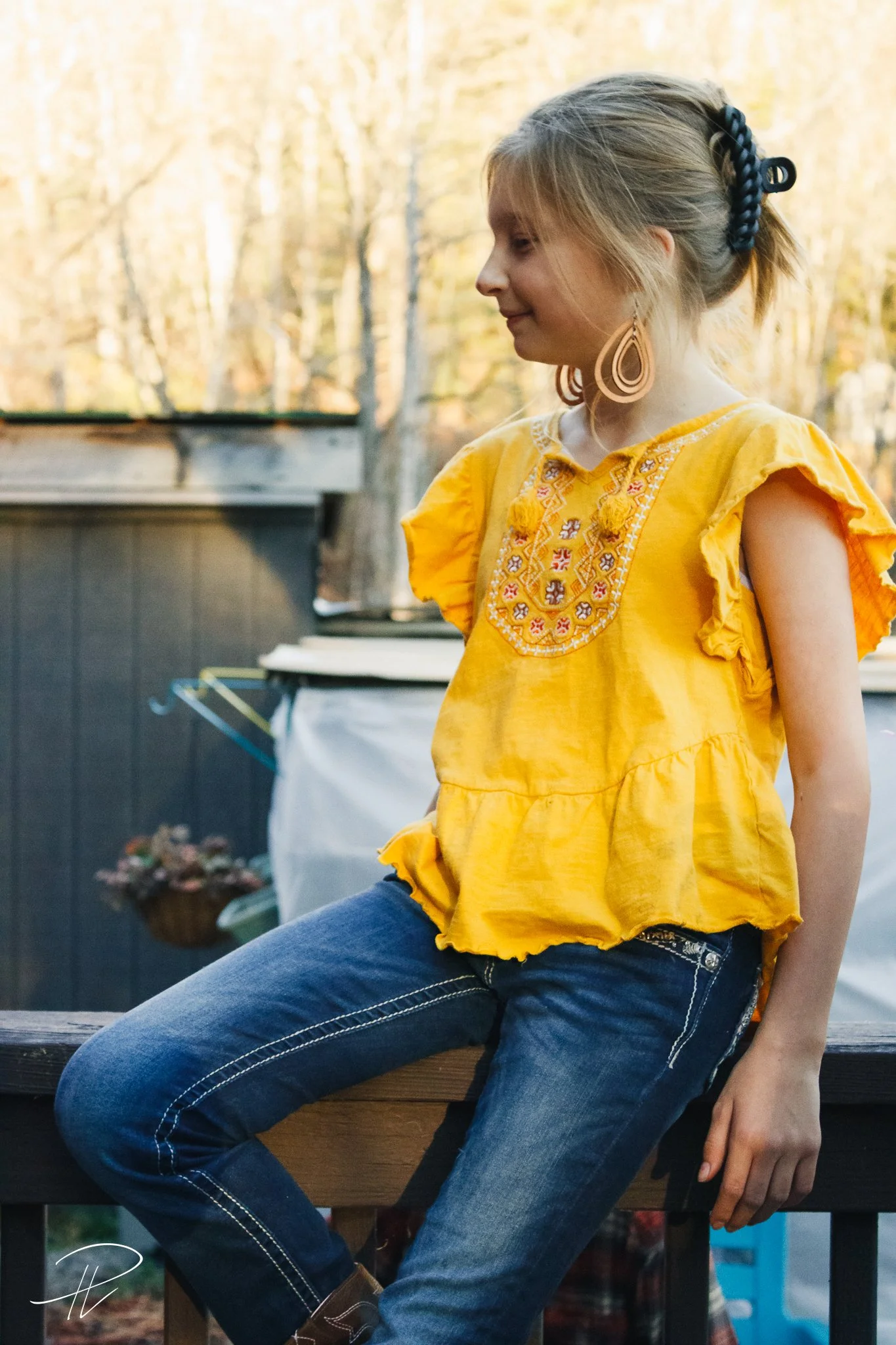 A young girl with blonde hair in a braid, wearing a yellow embroidered blouse with ruffled sleeves and blue jeans, sitting outdoors during the daytime.