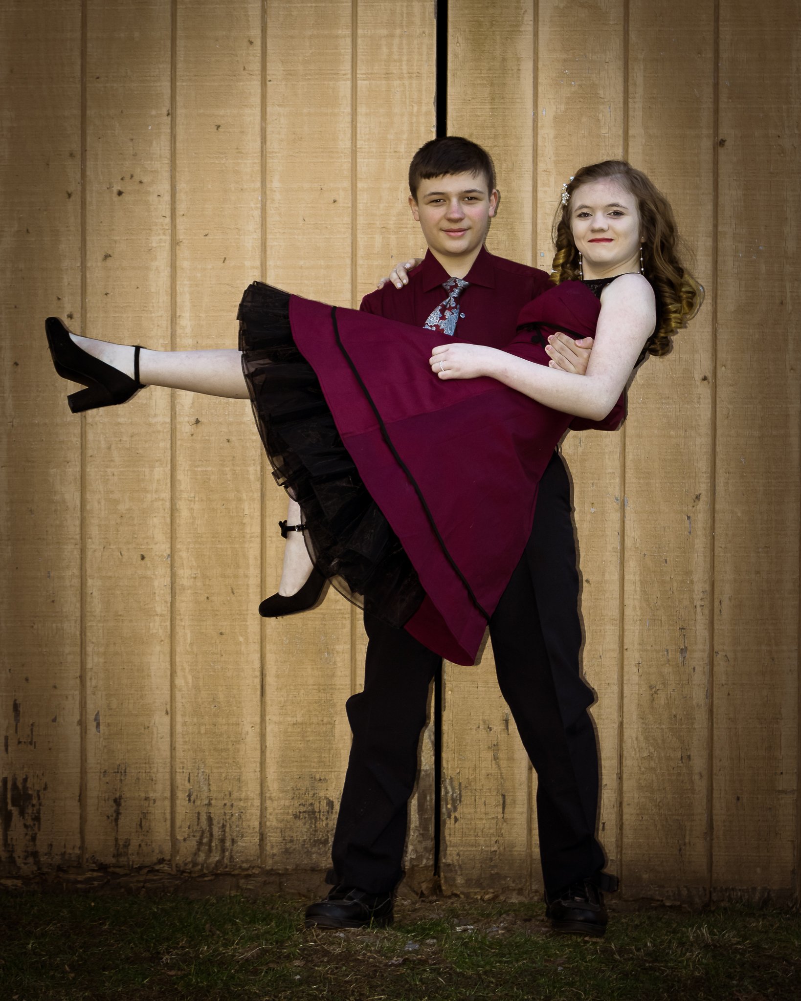 A young man in formal attire holding a young girl in a burgundy dress and black shoes in front of a wooden fence.