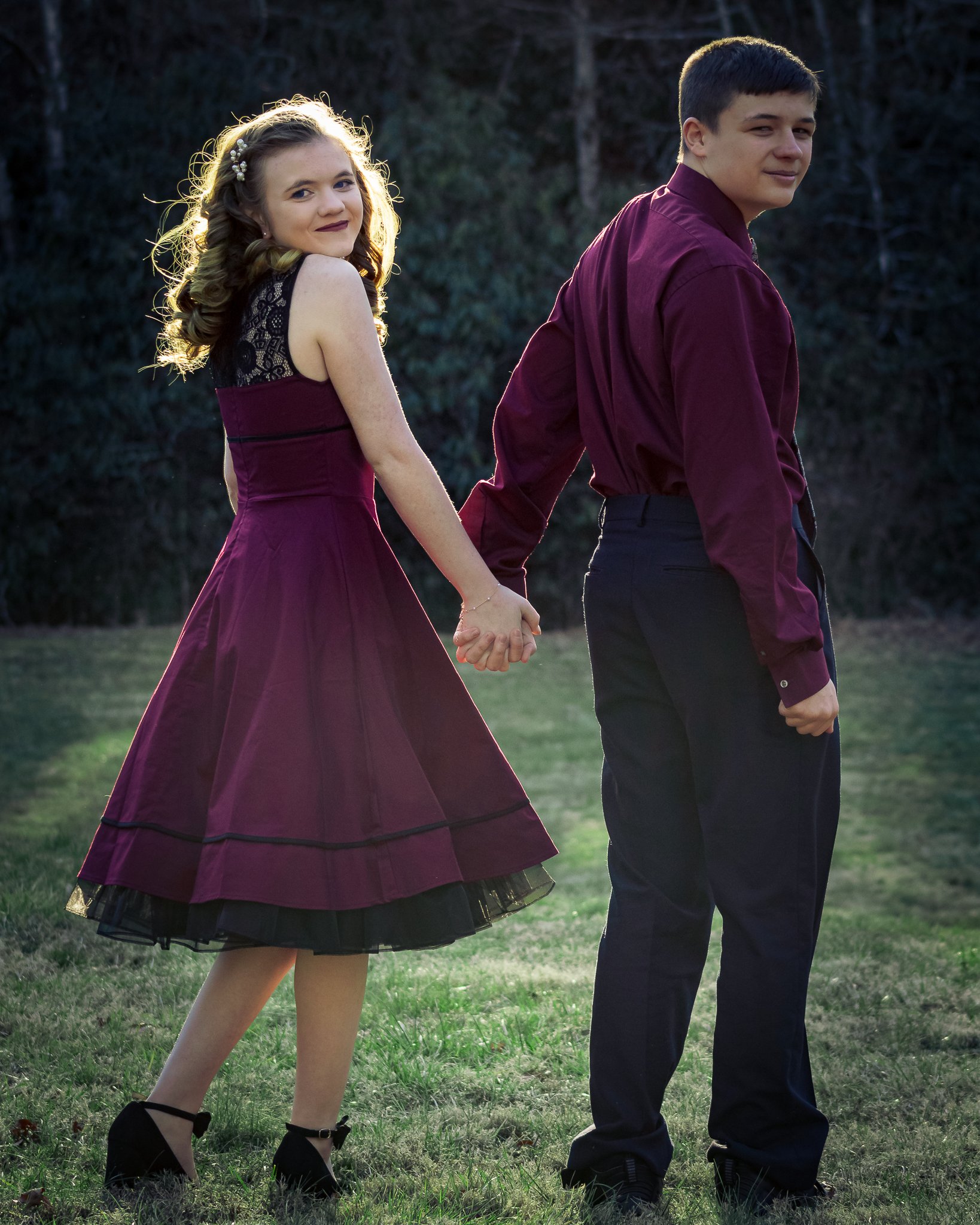 A young girl and boy holding hands and walking outdoors on grass during sunset, both dressed in formal attire, smiling at the camera.