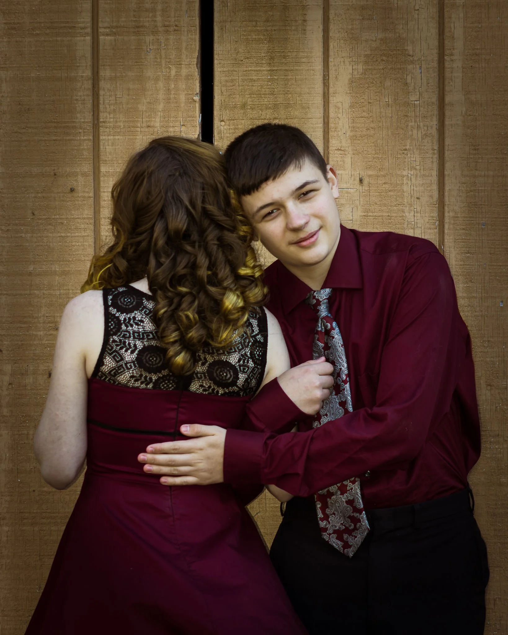 A teenage boy and girl in formal attire standing close against a wooden backdrop. The girl has curly hair and is wearing a maroon dress with black lace details, while the boy has short dark hair and is in a maroon shirt with a patterned tie.