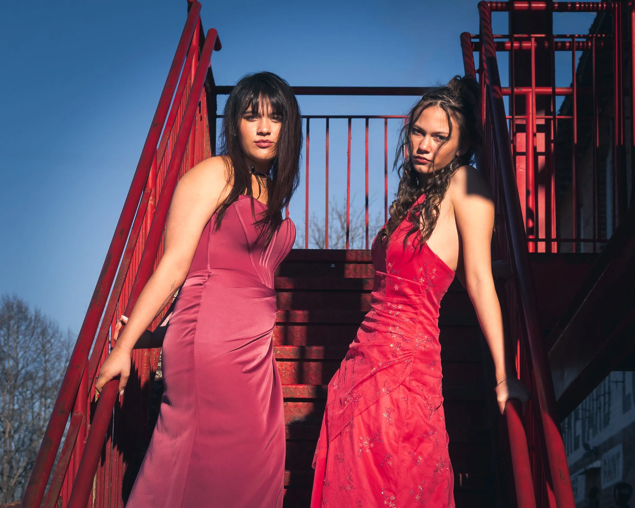 Two women standing on a red outdoor staircase, wearing red and pink dresses, with a blue sky background.