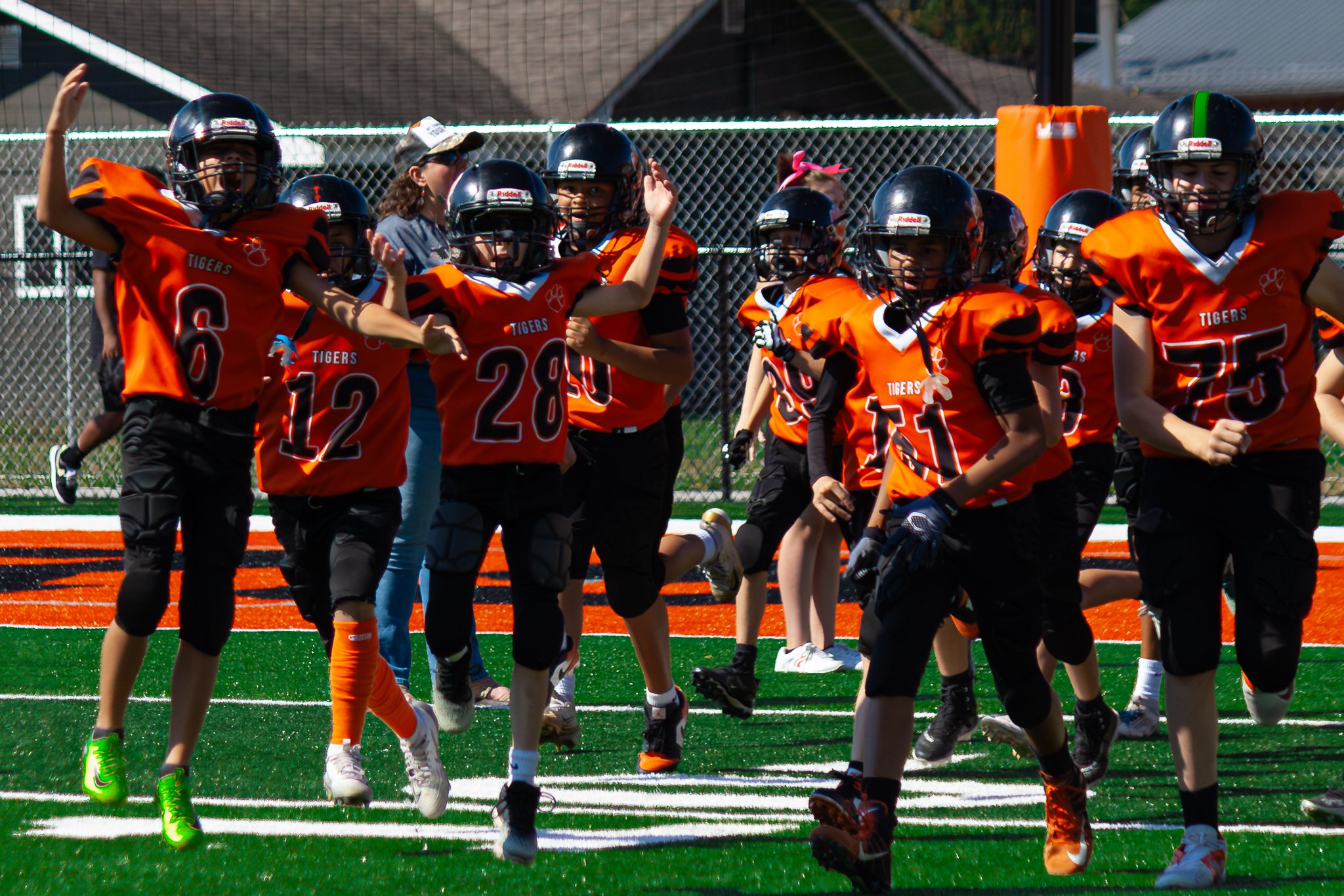 Children in orange and black football uniforms celebrating on a football field with a chain-link fence in the background.