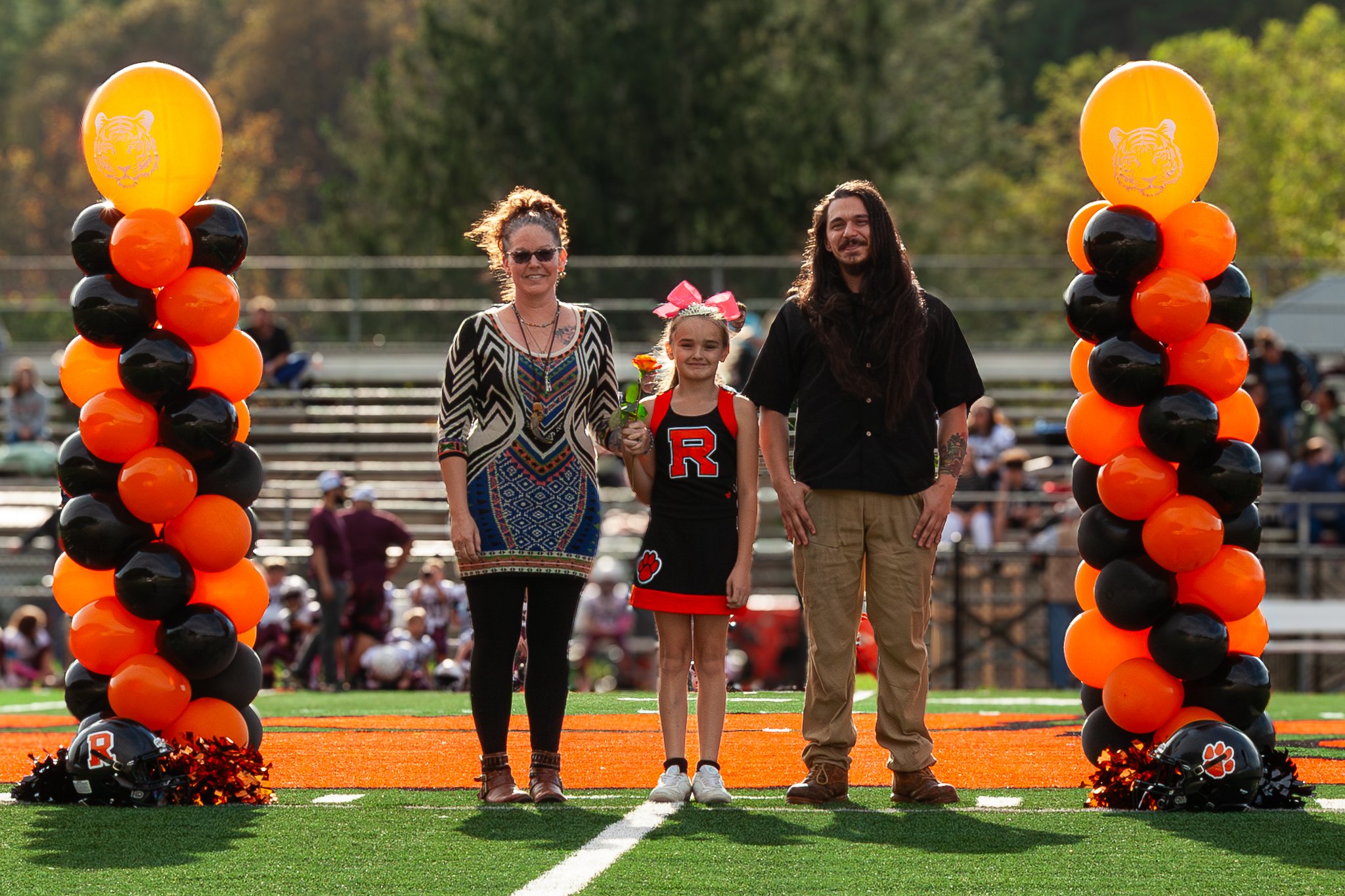 A girl in a cheerleading uniform standing between two adults on a sports field, with balloon columns in orange and black on either side, during daytime.
