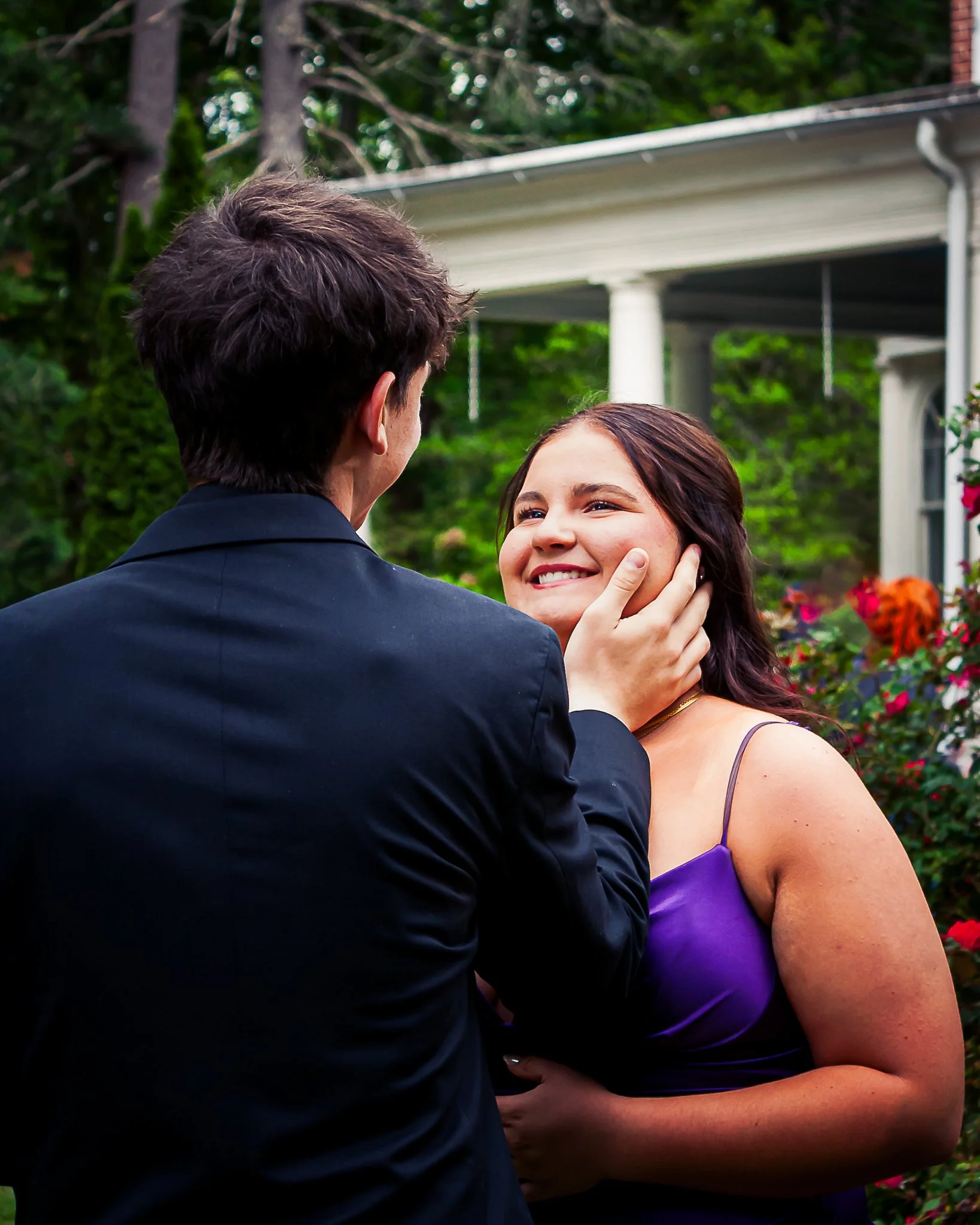 A man in a dark suit faces a woman in a purple dress, holding her face gently and smiling at her outdoors near a white house with a porch and lush greenery.