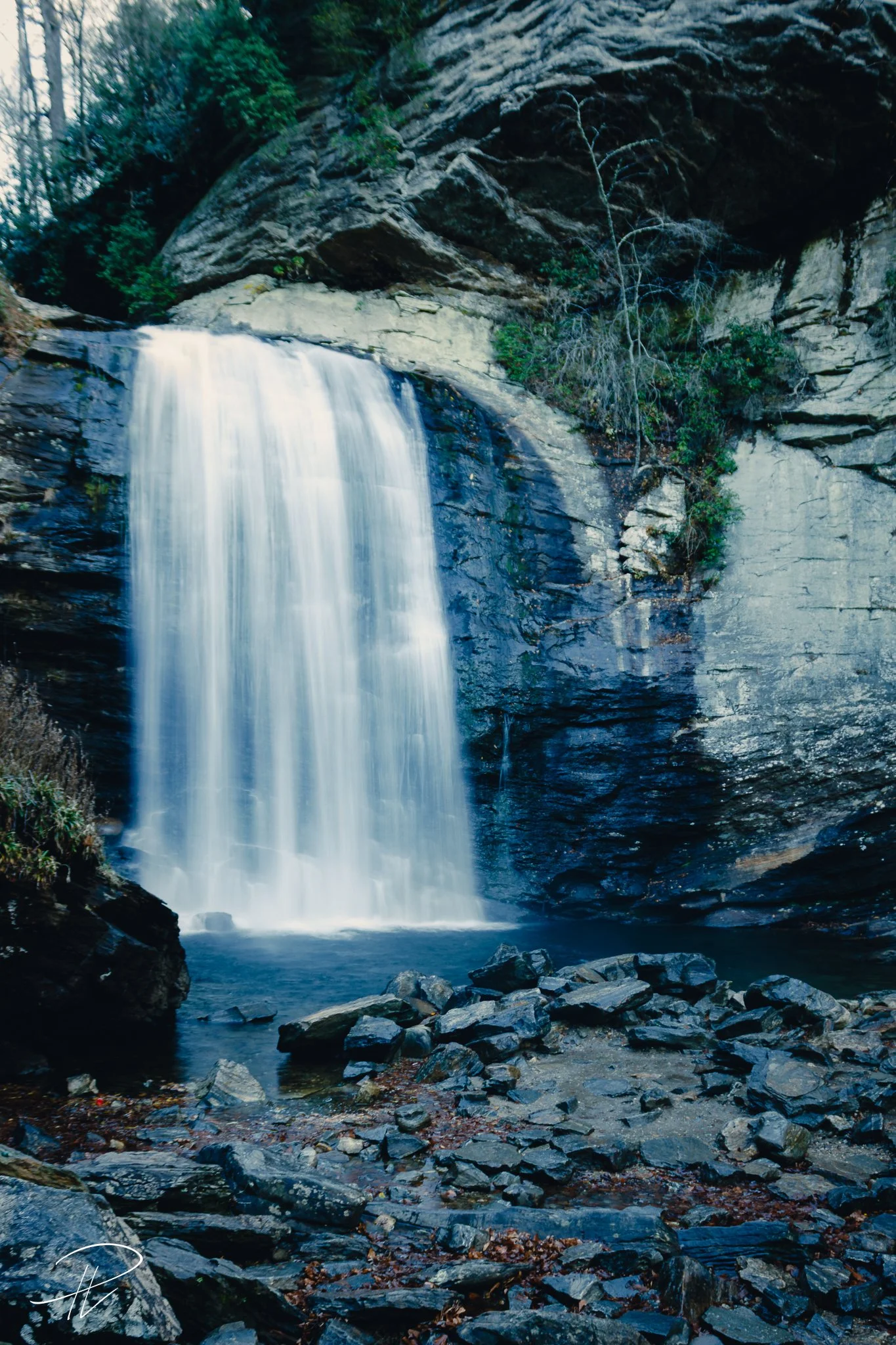 A scenic view of a waterfall cascading over rock cliffs into a rocky pool below, surrounded by trees and vegetation.