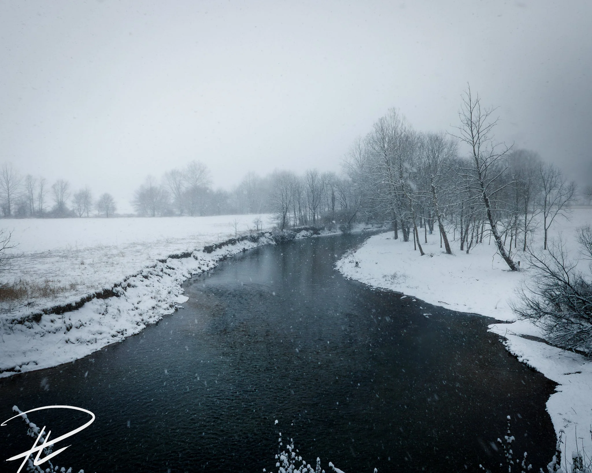 Snow-covered landscape with a river winding through the scene, trees with snow on their branches, and a foggy, overcast sky.