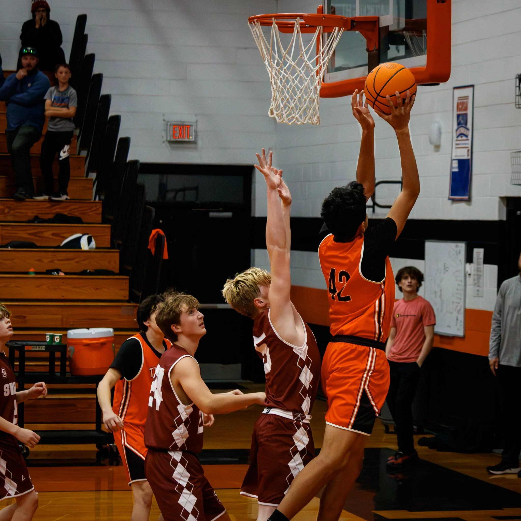 Young basketball player in orange jersey number 42 making a shot at the hoop while others in brown jerseys attempt to block in a gymnasium.