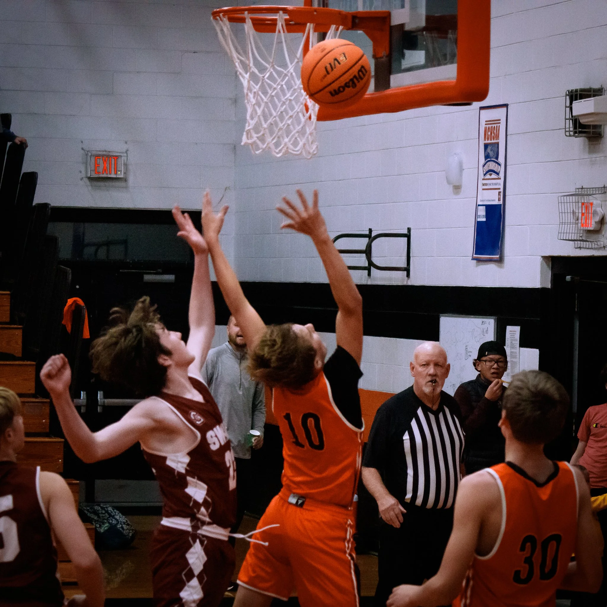 Two basketball players are jumping to reach the basketball near the hoop during a game in a gymnasium, with a referee and other players observing.