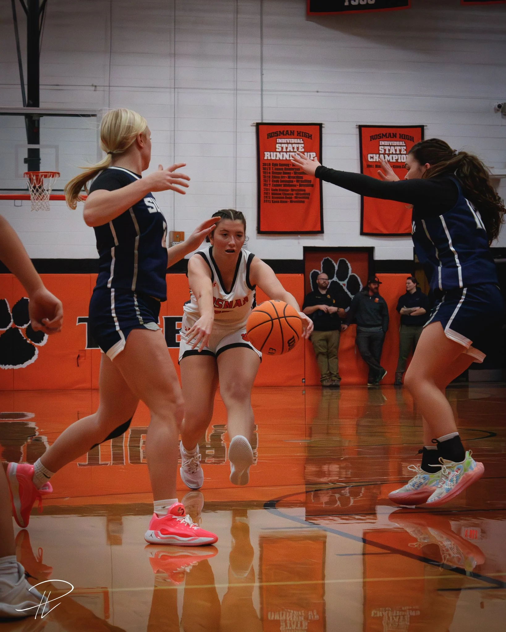 A women's basketball game with player holding ball while being guarded by two defenders in a gymnasium with orange walls and banners.
