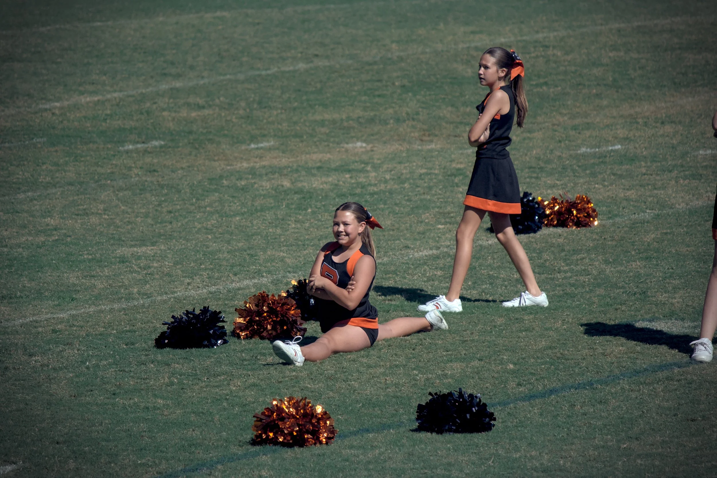 Two young cheerleaders in black and orange uniforms with pom-poms on a football field, one sitting on the ground and smiling with pom-poms, the other standing with arms crossed.