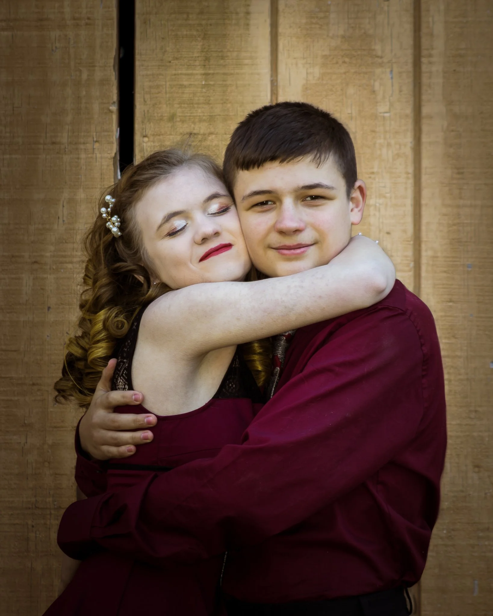A girl with curly hair and makeup hugging a boy with short hair against a wooden background.