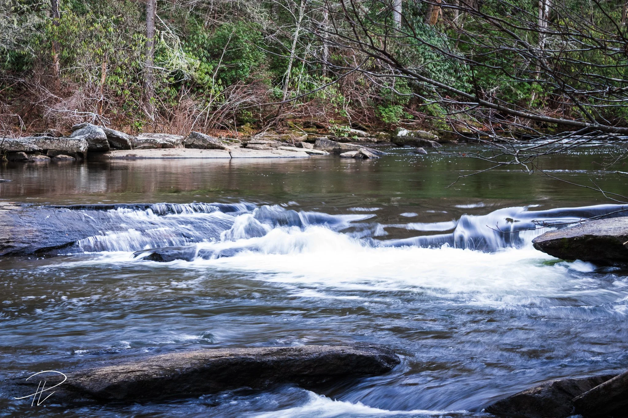 A peaceful river flows through a wooded area with rocks and trees along the banks.